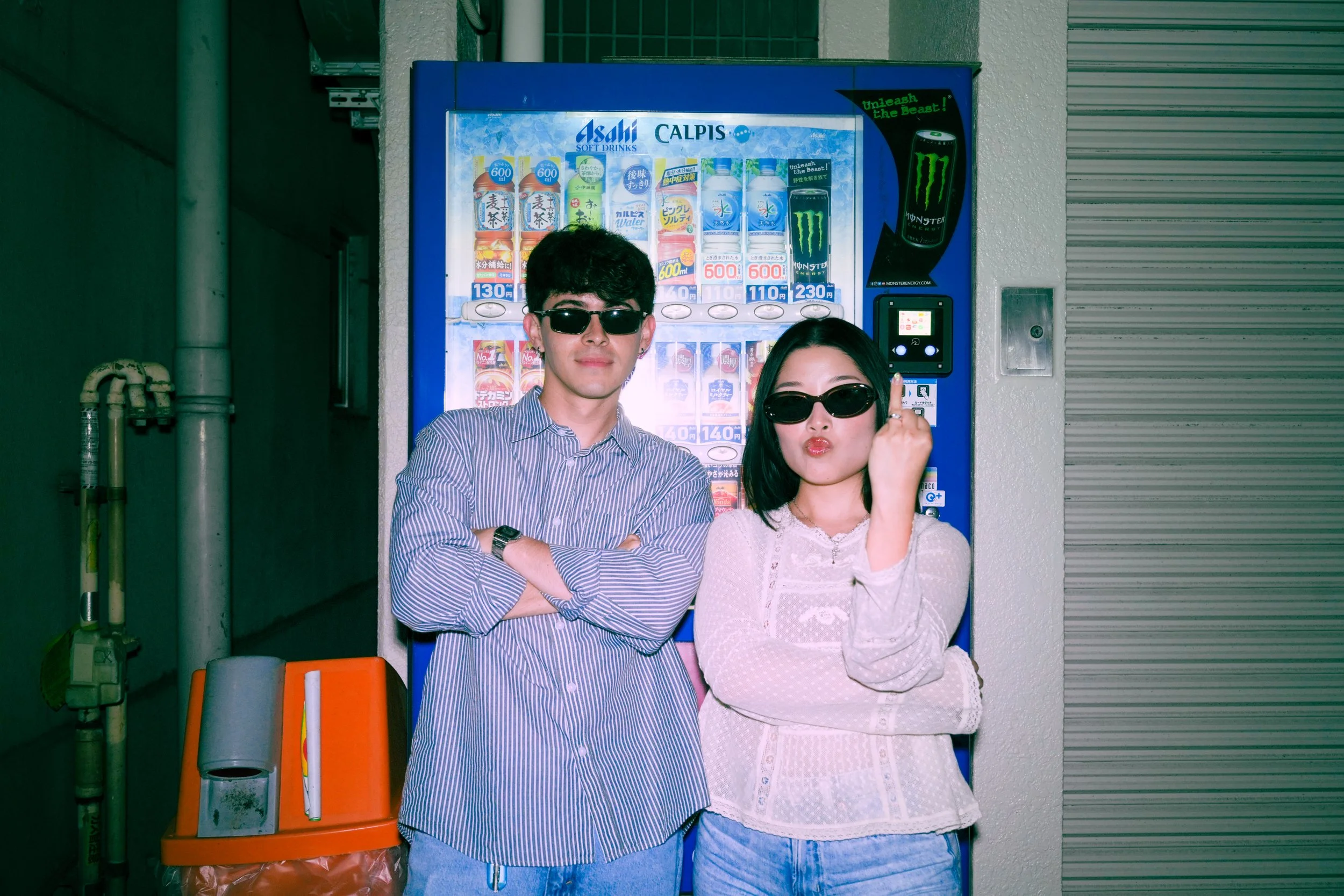 A young man and woman wearing sunglasses posing in front of a vending machine. The man is crossing his arms and the woman is holding her finger to her temple.
