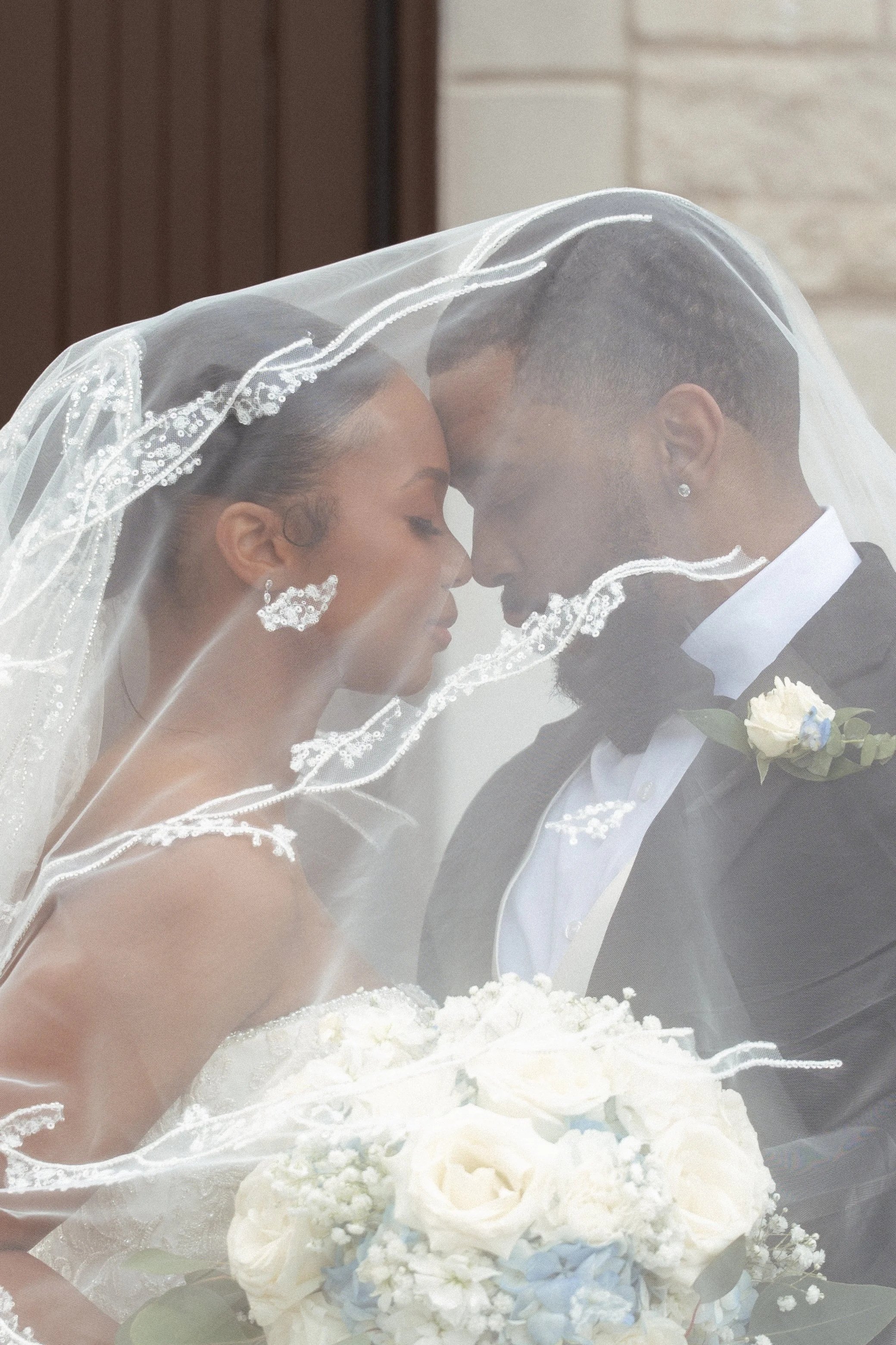 A bride and groom with their foreheads touching, partially covered by a sheer wedding veil, standing behind a bouquet of white flowers.