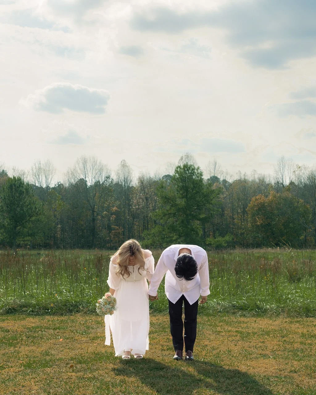 A bride and groom bowing their heads together in a field, holding hands, during an outdoor wedding ceremony in a grassy area with trees and a cloudy sky in the background.