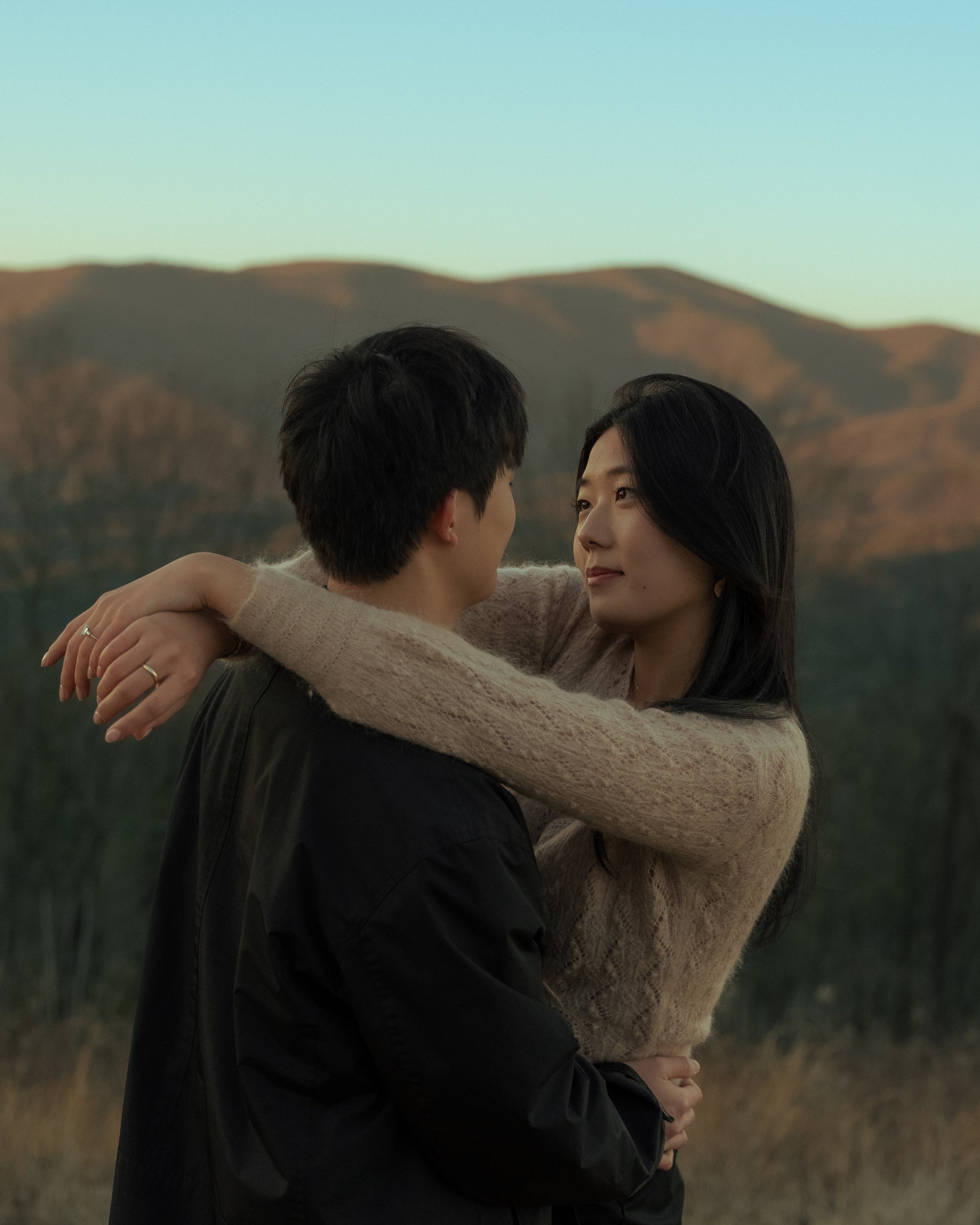 A young couple embracing outdoors at dusk, with mountains in the background.