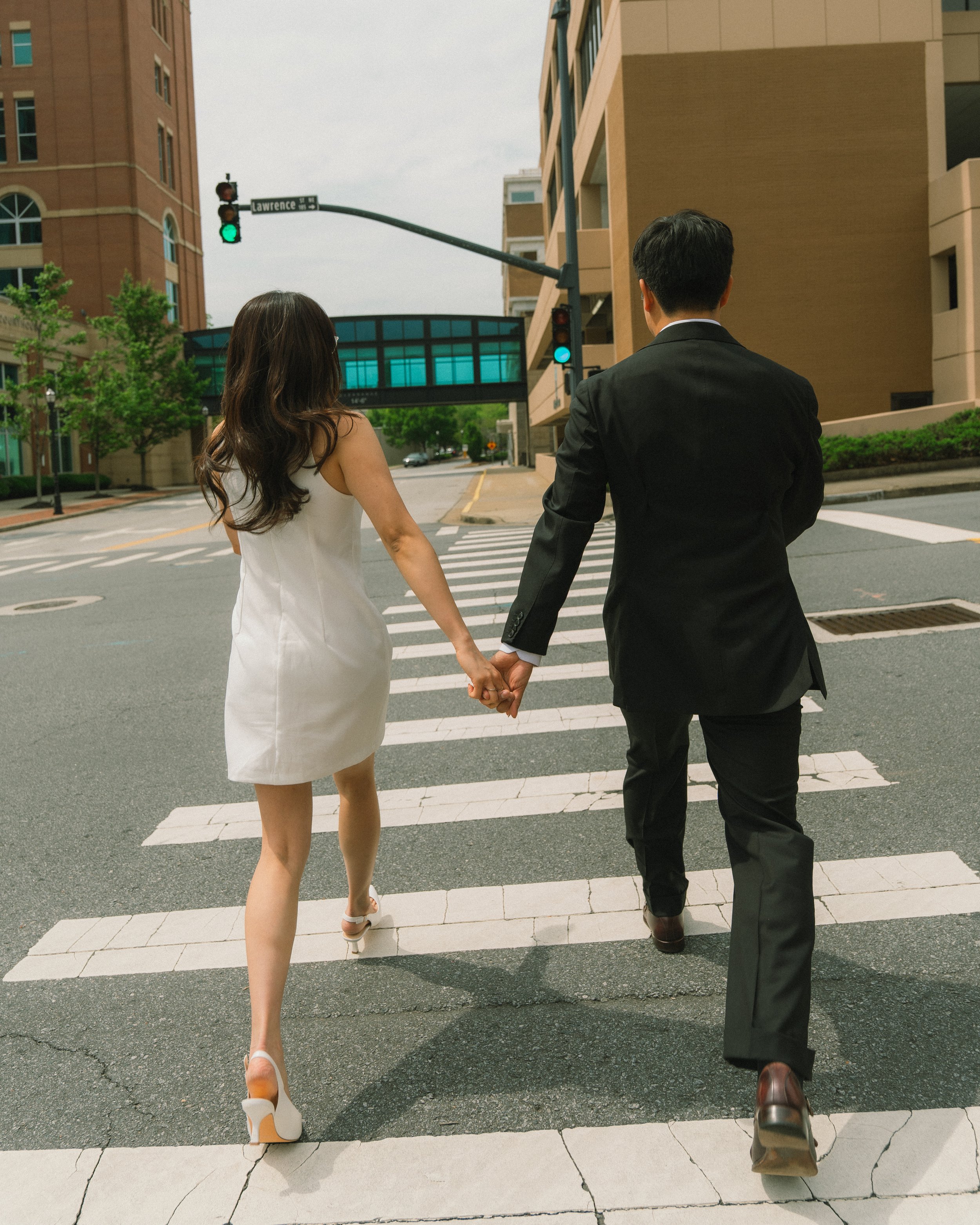 A couple holding hands crossing a city street at a crosswalk, with tall buildings and traffic signals visible.