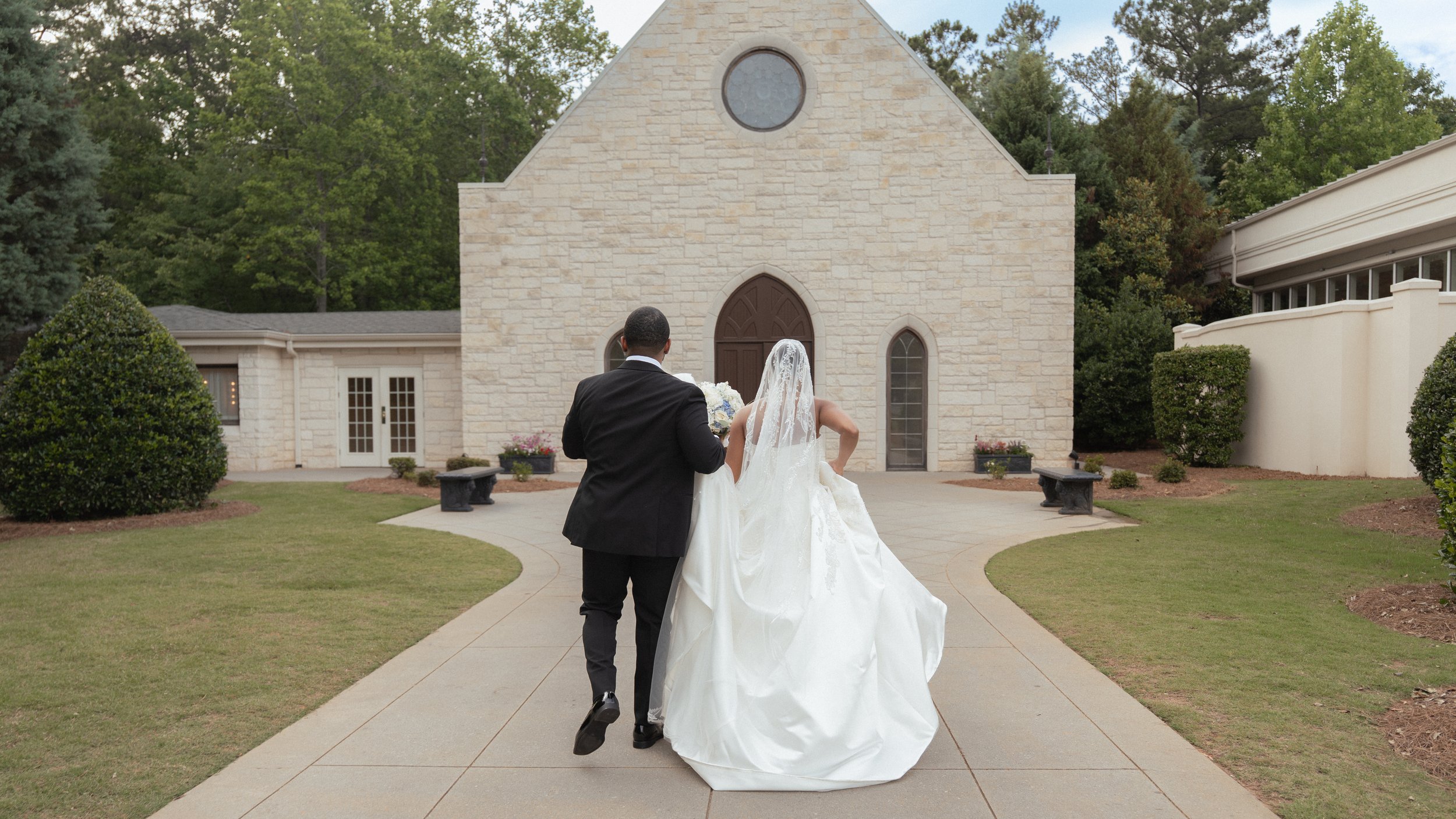 A bride and groom walking towards a small stone chapel for their wedding ceremony on a paved pathway surrounded by grass and bushes.