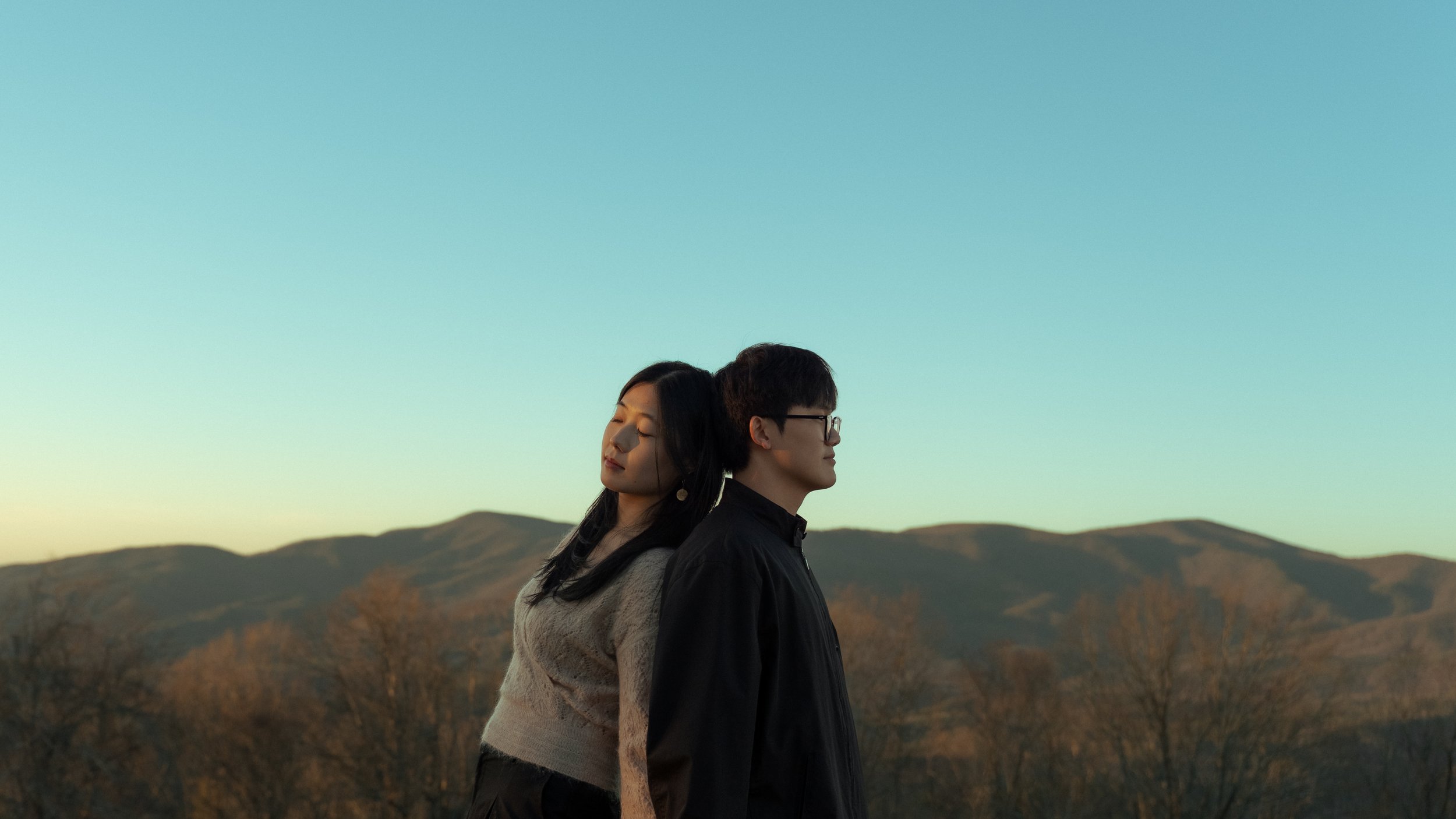 A man and woman stand back-to-back outdoors during sunset with mountains in the background, the woman with eyes closed and the man wearing glasses.
