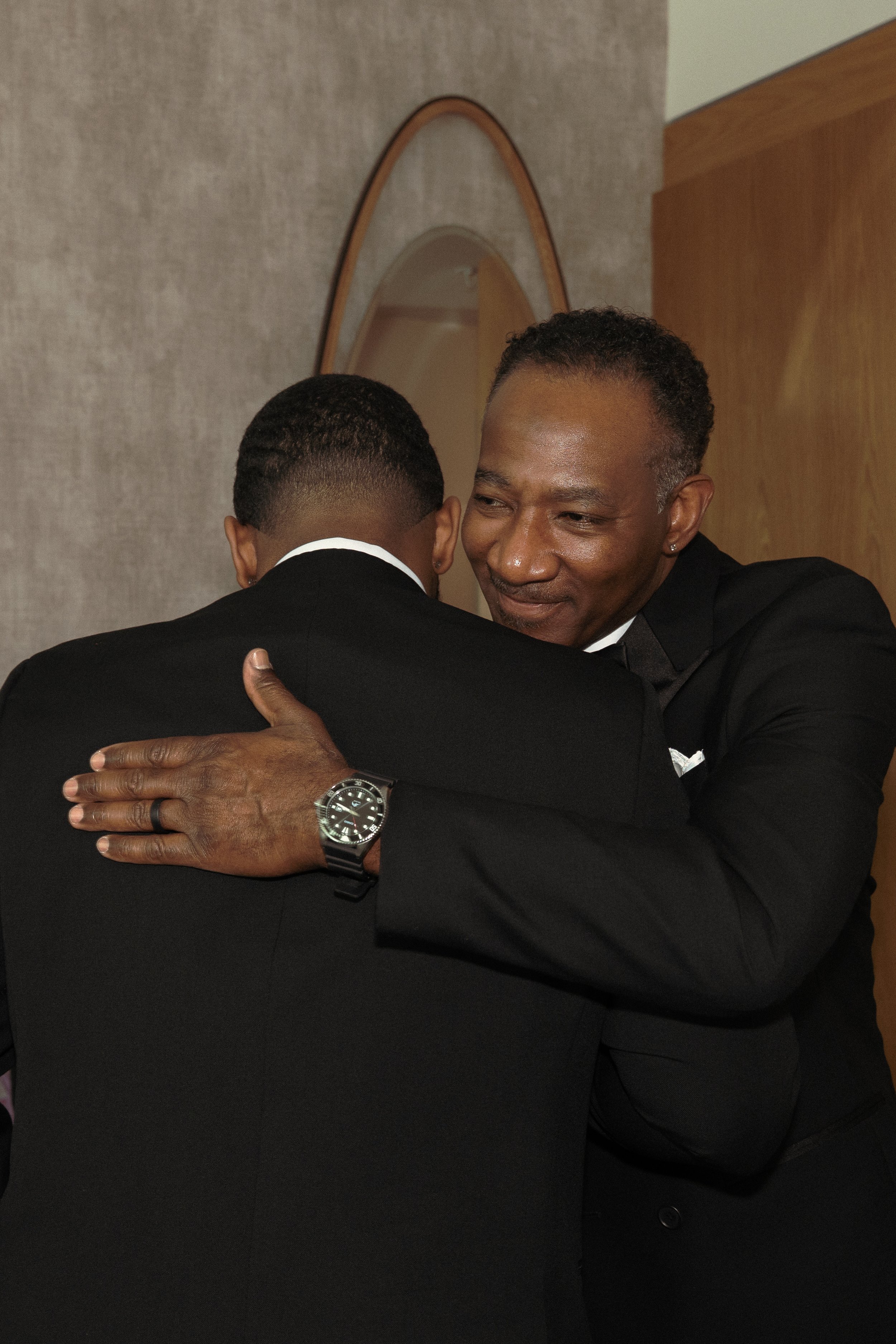 Two men in black suits embracing in a hug, one with a watch on his wrist and an earring, in a room with beige walls and a mirror.