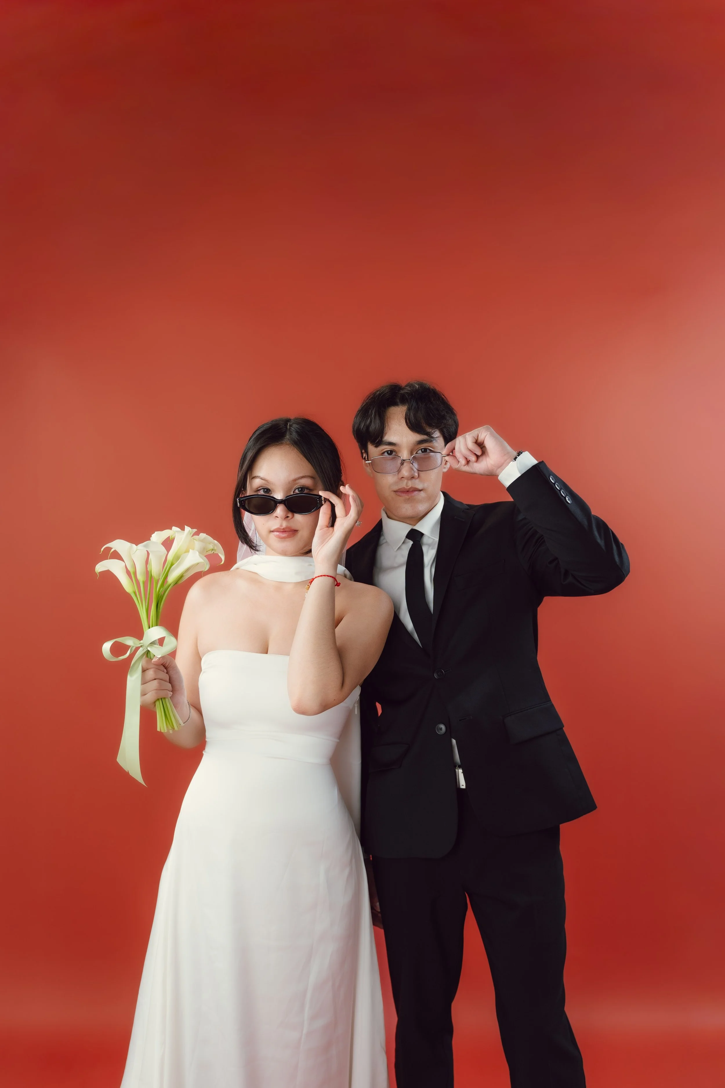 A man and woman dressed in formal attire pose against a red-orange background. The woman is in a strapless white dress holding a bouquet of white lilies with a yellow ribbon, and wearing black sunglasses. The man wears a black suit with a white shirt and black tie, and is adjusting his glasses with his right hand. They look directly at the camera.