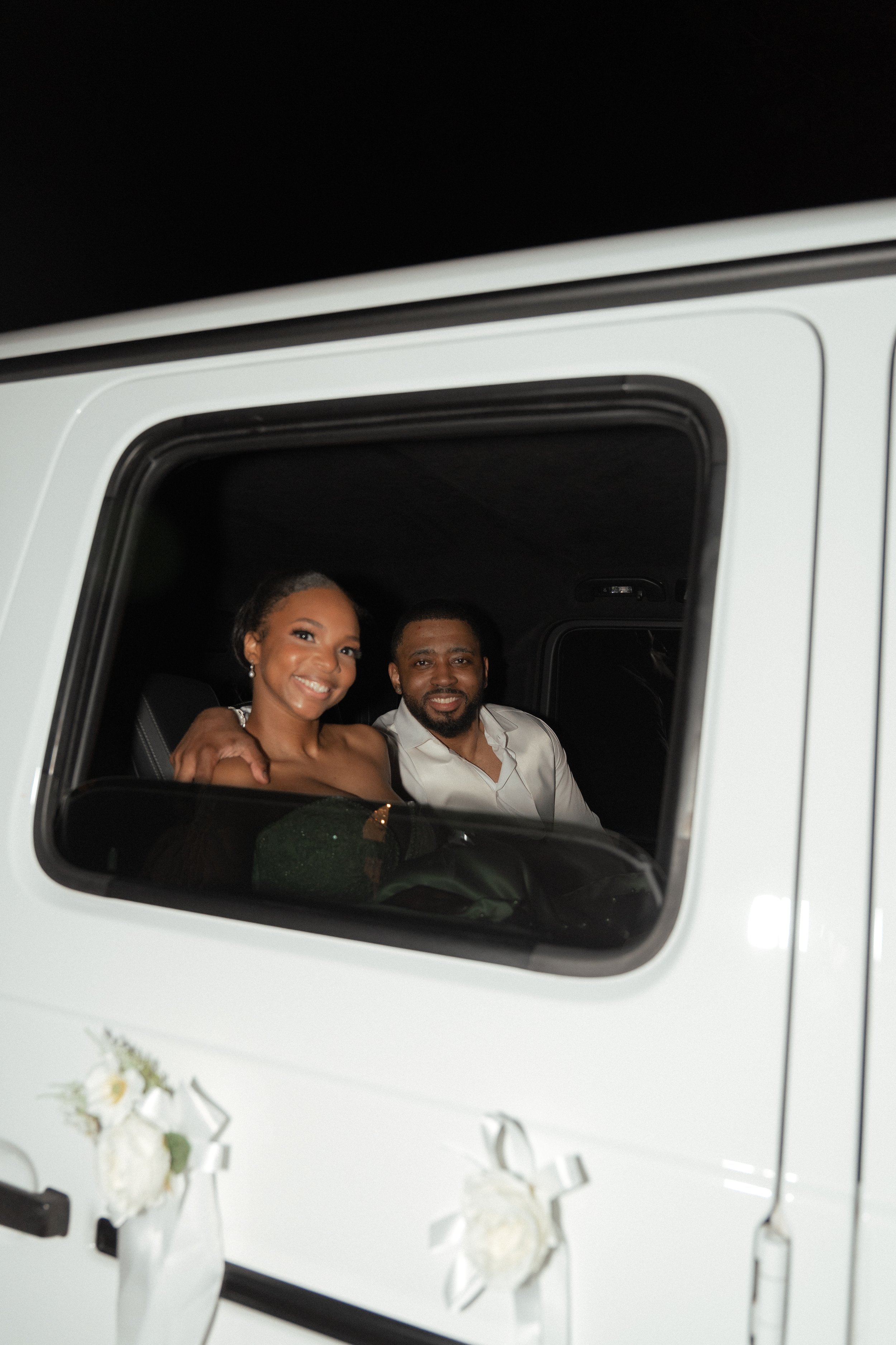 A smiling couple sitting inside a white vehicle, possibly a classic or vintage car, with the woman on the left and the man on the right, both with dark hair, dressed nicely, on a night or dark background.