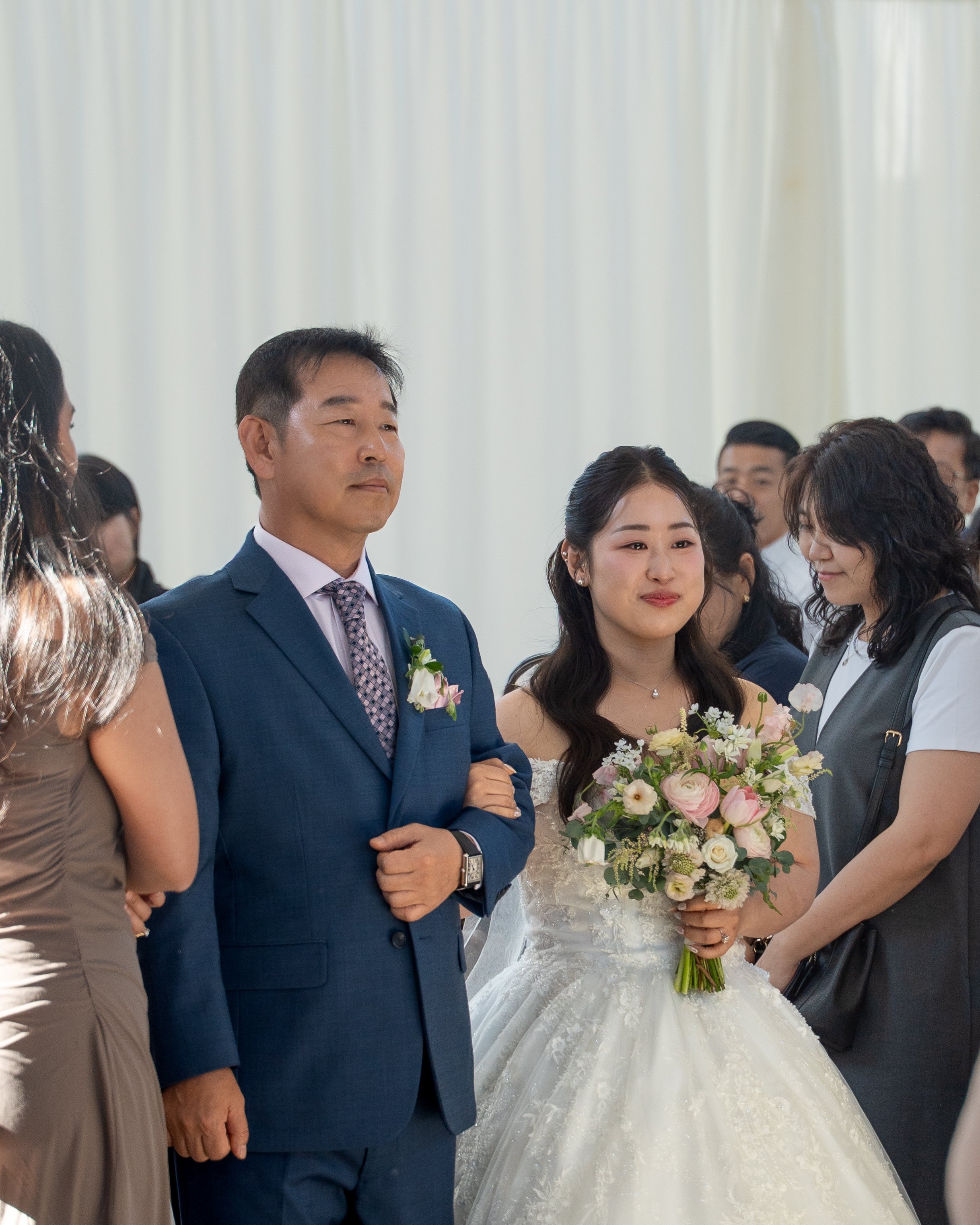 A bride in a wedding dress holding a bouquet of flowers being walked down the aisle by her father in a blue suit during a wedding ceremony. They are surrounded by wedding guests dressed in formal attire, indoors with a light-colored curtain backdrop.