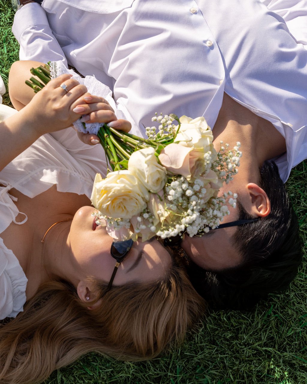A couple lying on grass, holding a bouquet of flowers between them, with their faces close together, one wearing sunglasses and the other with their eyes closed.