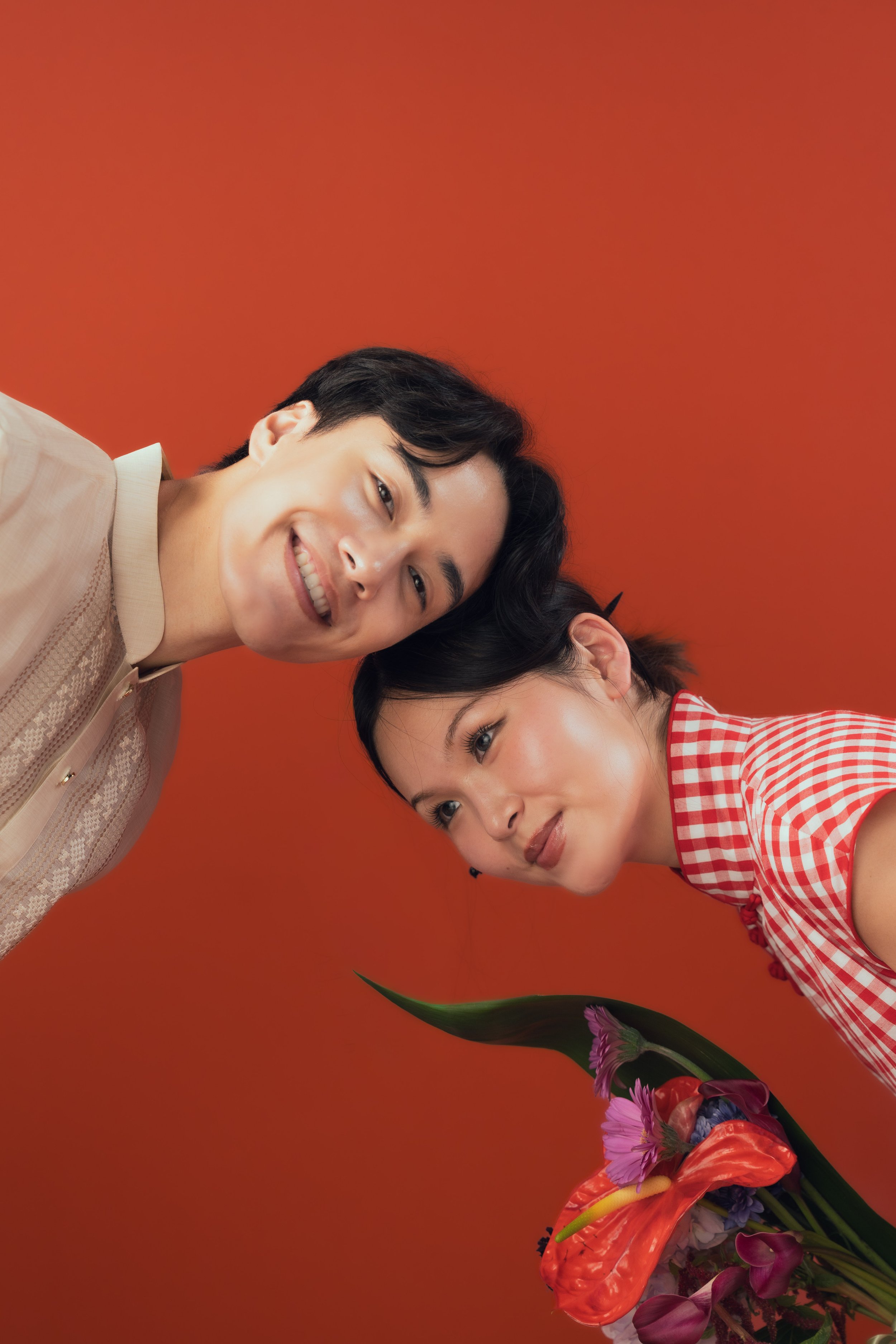A young man with black hair and a young woman with black hair and bangs are smiling and looking at the camera against an orange background, with a bouquet of colorful flowers in the lower right corner.