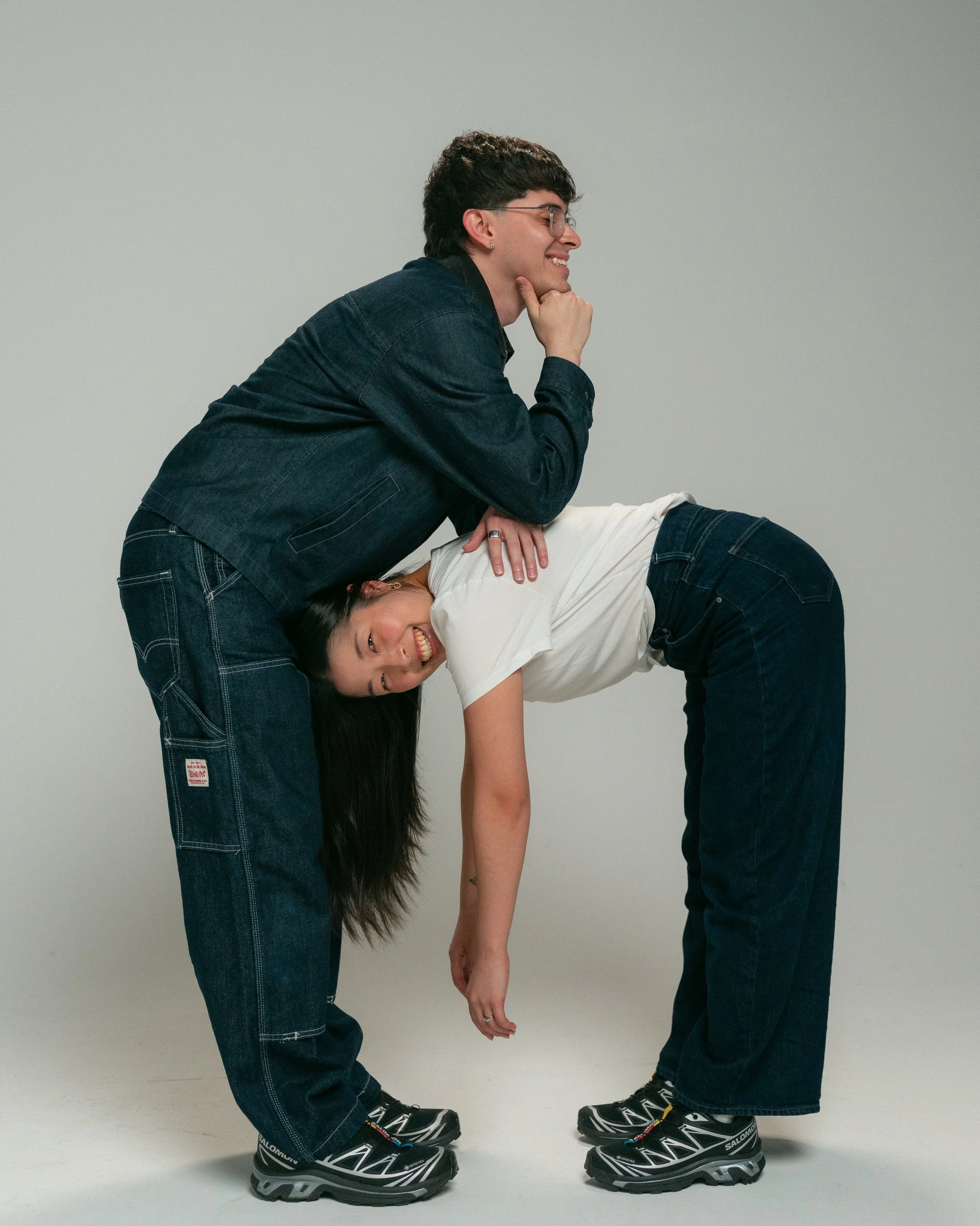 Two people wearing black Salomon hiking shoes are holding a third person in a yoga pose. The person being held is smiling and bending forward with their hands and one foot on the ground, the other foot and hands are hanging downward. The person on top is smiling and leaning their chin on their hand.