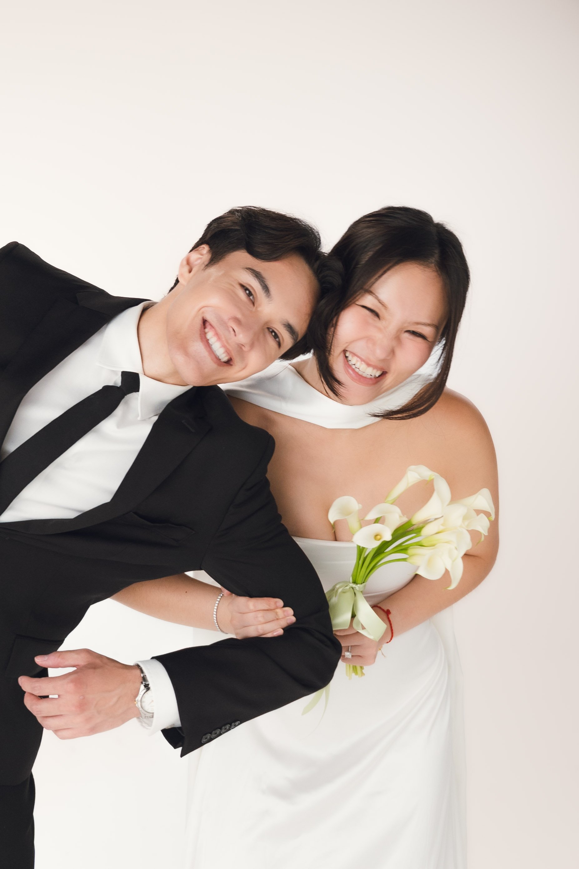 A happy bride and groom posing together, the groom in a black tuxedo and the bride in a white wedding dress holding a bouquet of calla lilies.