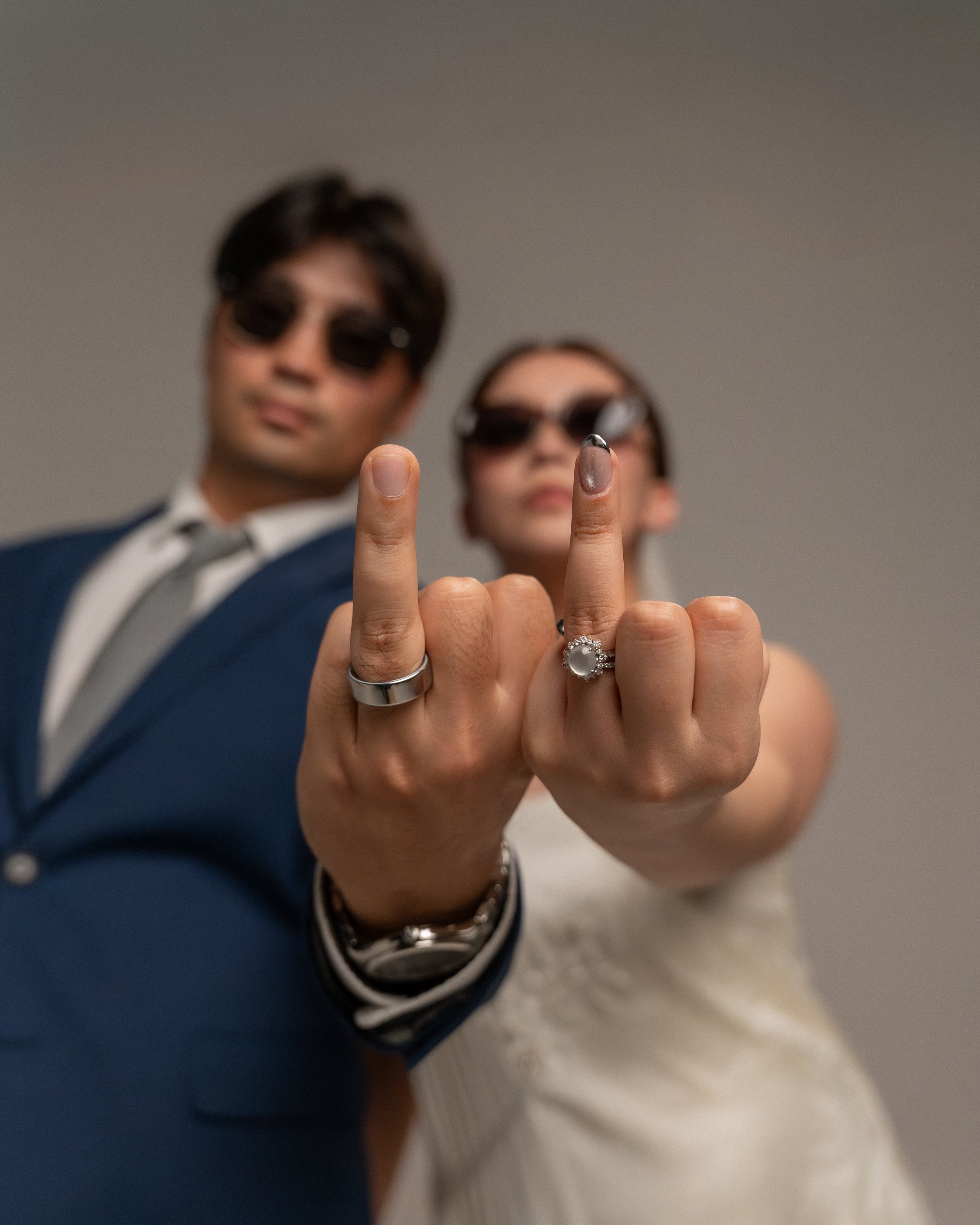 Two people, a man and a woman, are blurred in the background wearing sunglasses, while their fingers are in sharp focus in the foreground showing wedding rings and painted nails. The man is dressed in a suit, and the woman is in a dress.
