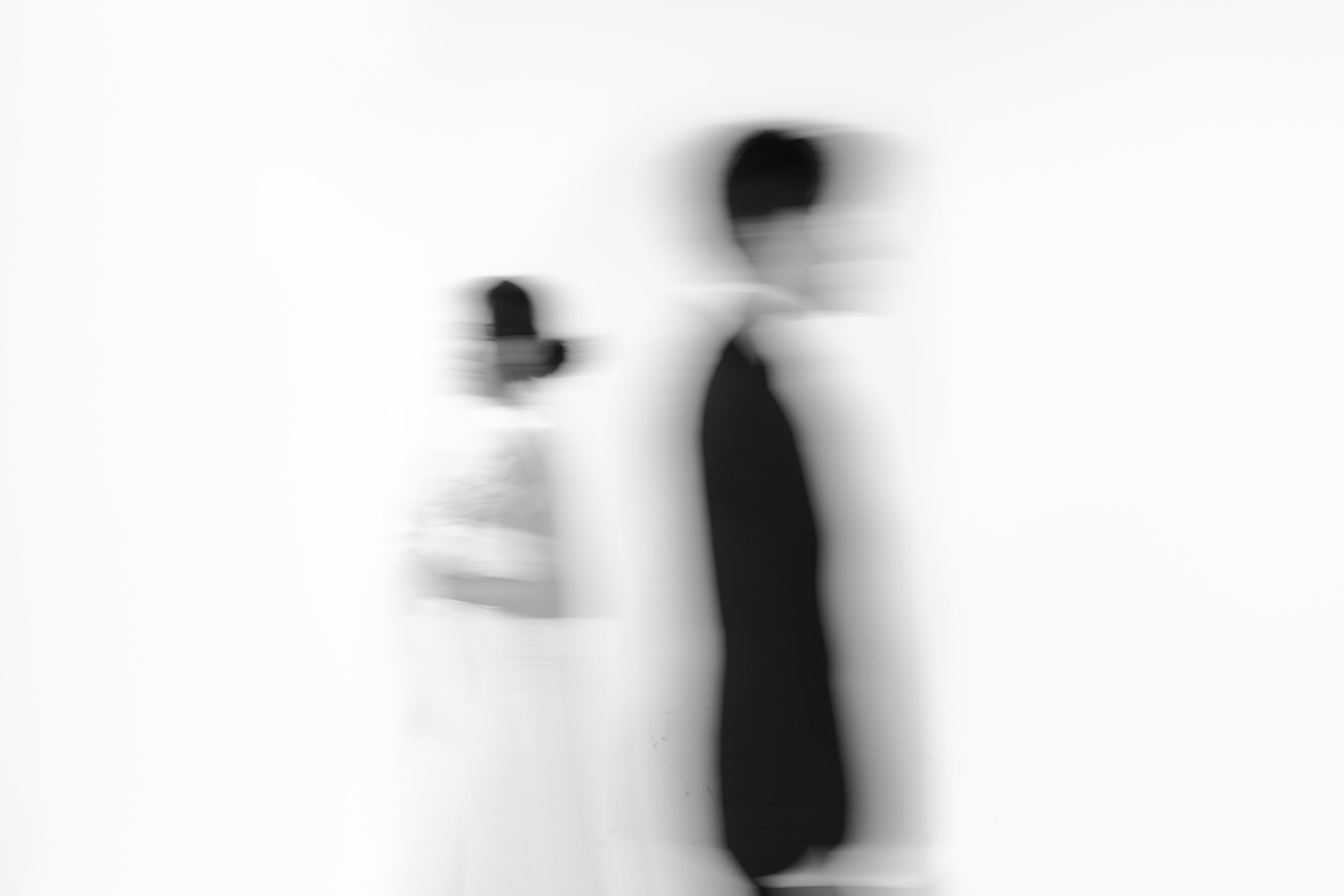 A blurred grayscale image of a bride and groom in profile passing by each other, against a plain white background shot in a professional studio for an engagement session.