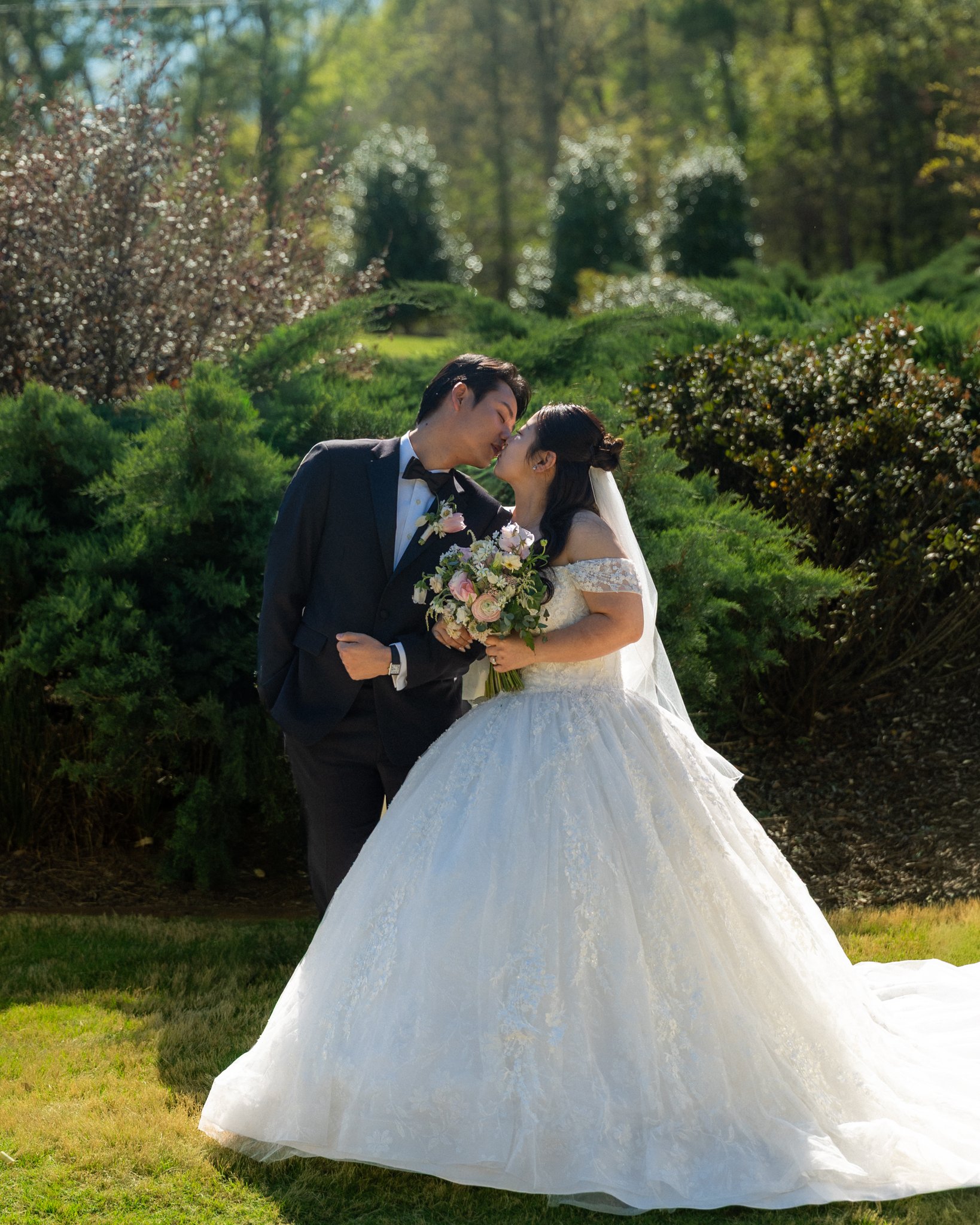 A bride and groom sharing a kiss in a garden, with the bride holding a bouquet of flowers, during a wedding photo shoot.