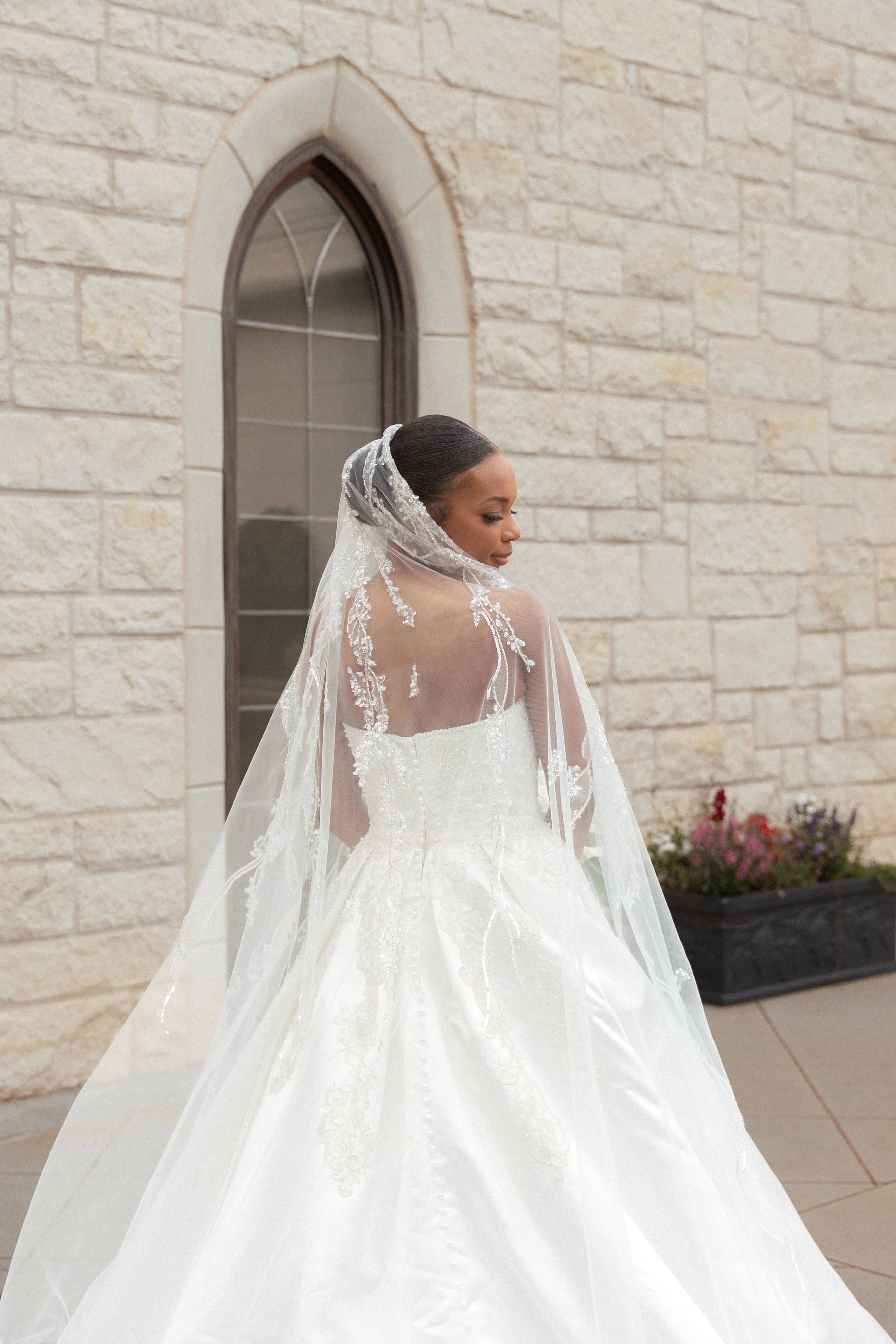 A bride in a white wedding gown with lace details, wearing a veil, standing outside near a beige brick building with a window, and a flower pot with colorful flowers.