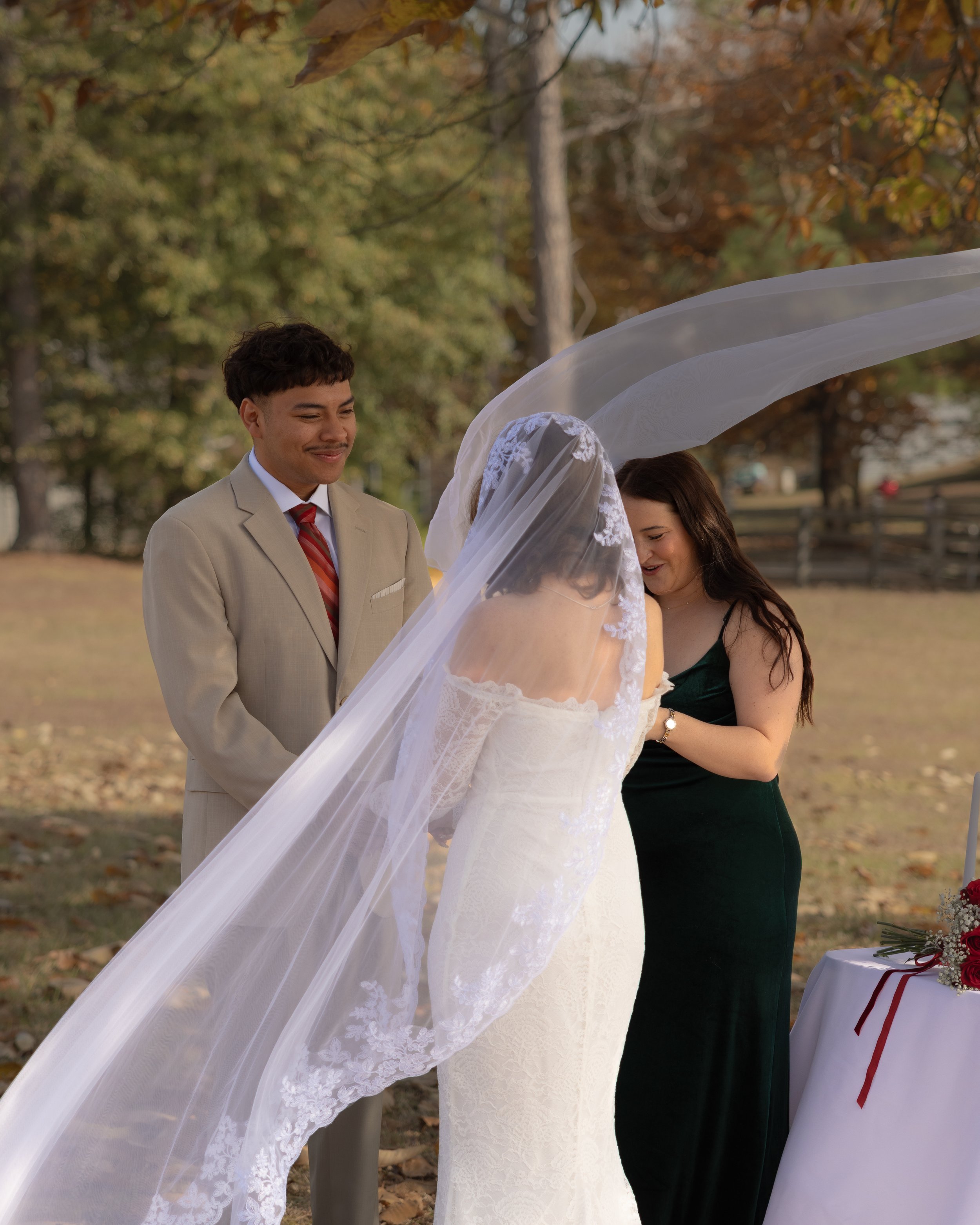 A wedding ceremony outdoors with three people, two women and one man. The bride is wearing a white lace wedding gown and veil, the groom is in a beige suit with a red striped tie, and a woman in a dark green dress is on the right. They are standing under a tree with fall foliage, with a table holding a bouquet of flowers visible to the side.