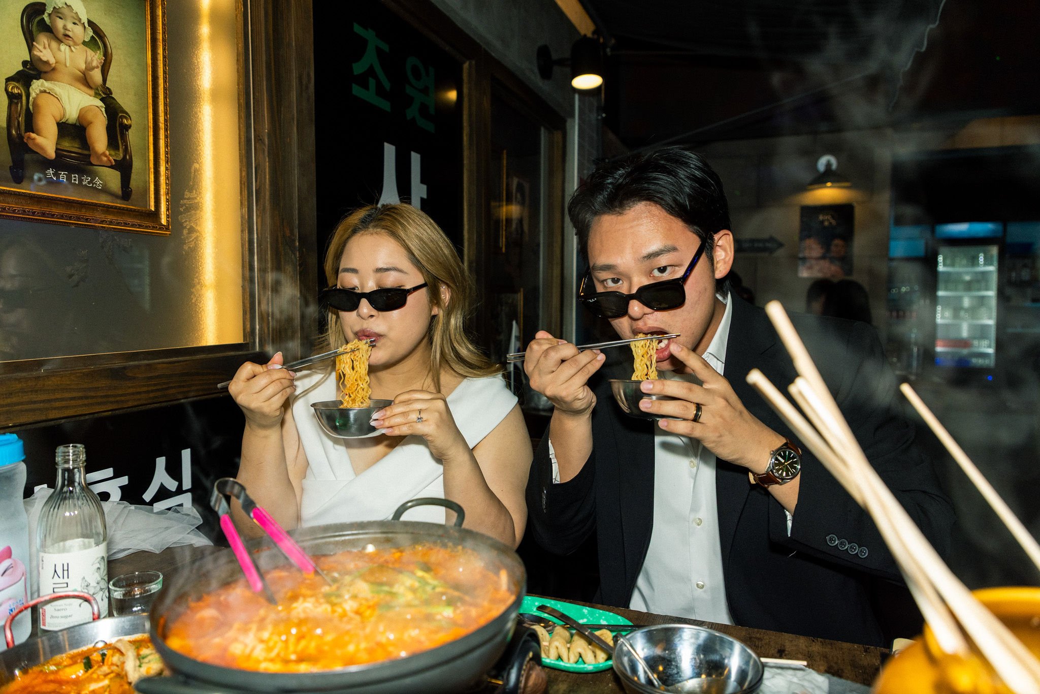 A man and woman wearing sunglasses are eating noodles at a restaurant table with various dishes and condiments, with a framed baby photo on the wall behind them.