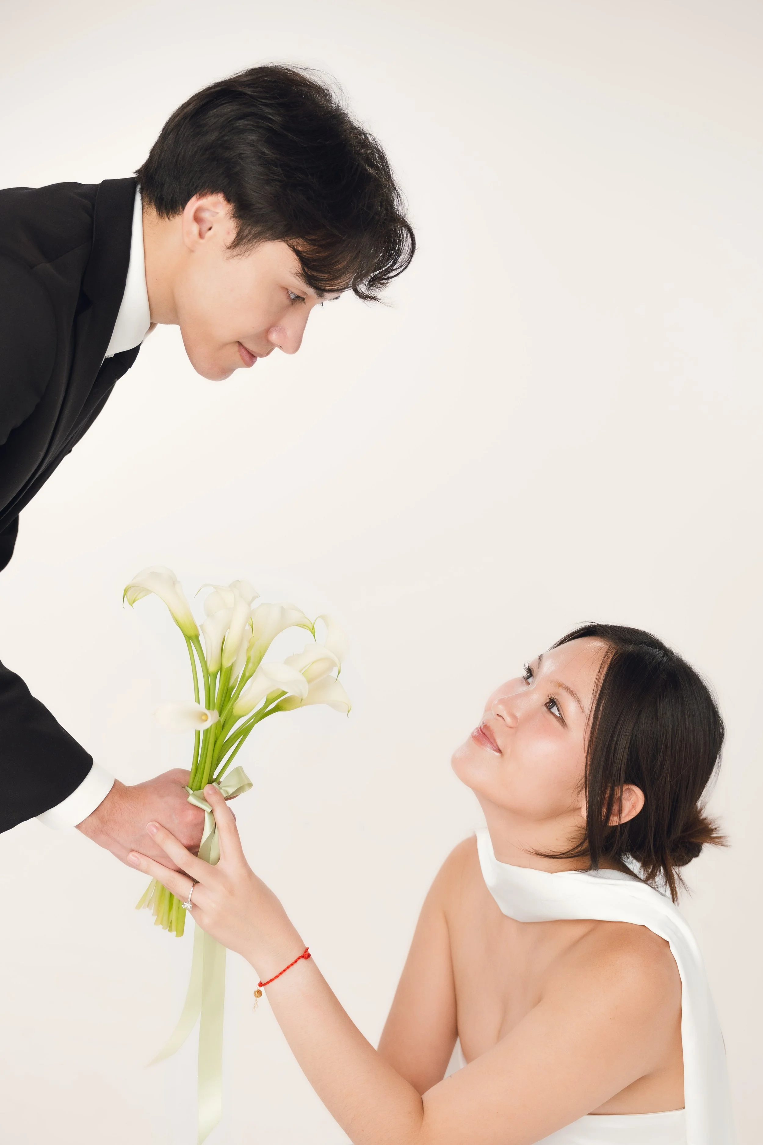 A man in a black suit and a woman with a white dress exchange a bouquet of white calla lilies in a bright studio with a plain background.