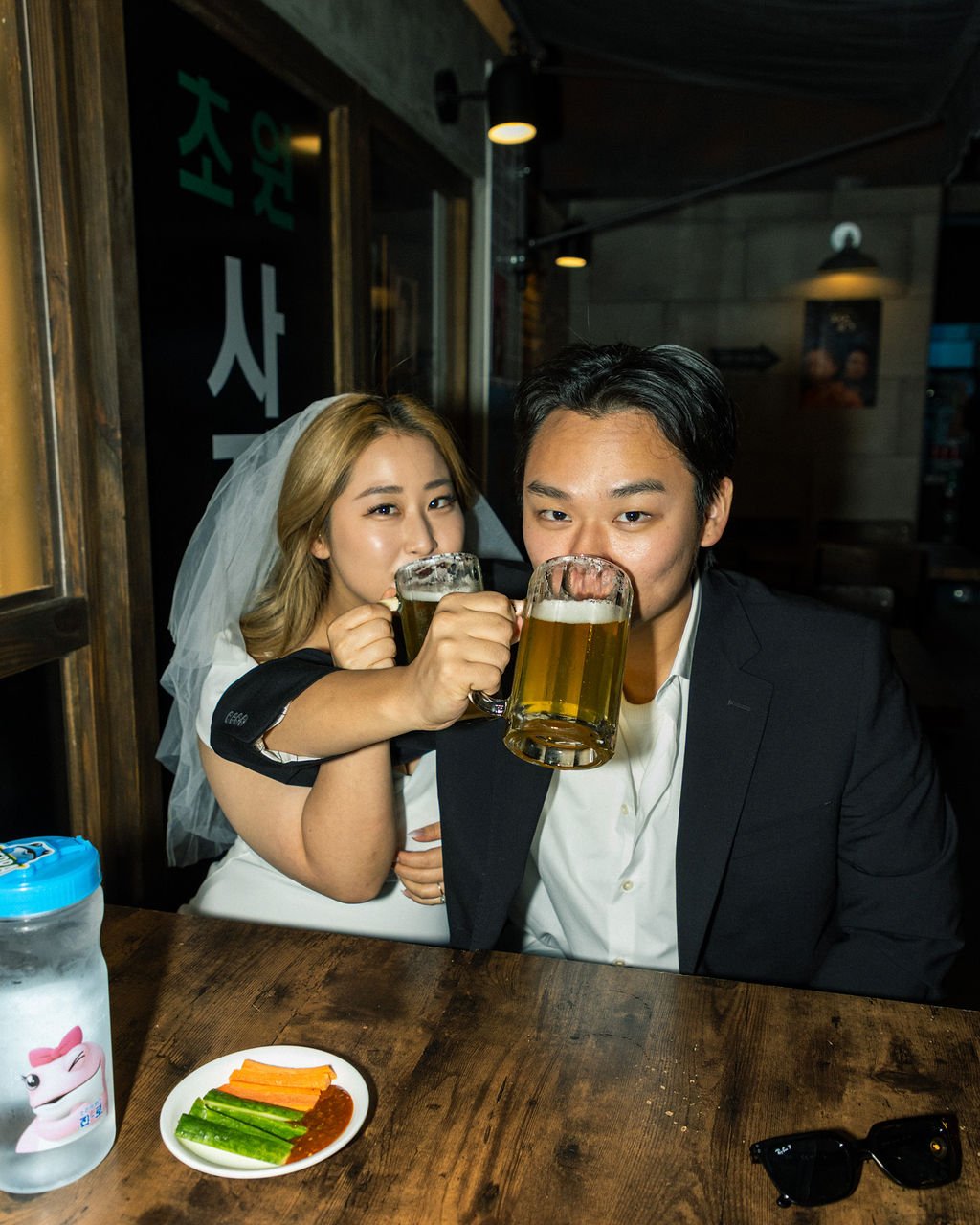 Two people, a woman wearing a wedding veil and a man in a suit, are sitting at a wooden table in a bar or restaurant. They are clinking glasses of beer and smiling. On the table, there is a small plate of vegetables and dip, a jar with a cartoon character on it, and a pair of black sunglasses.