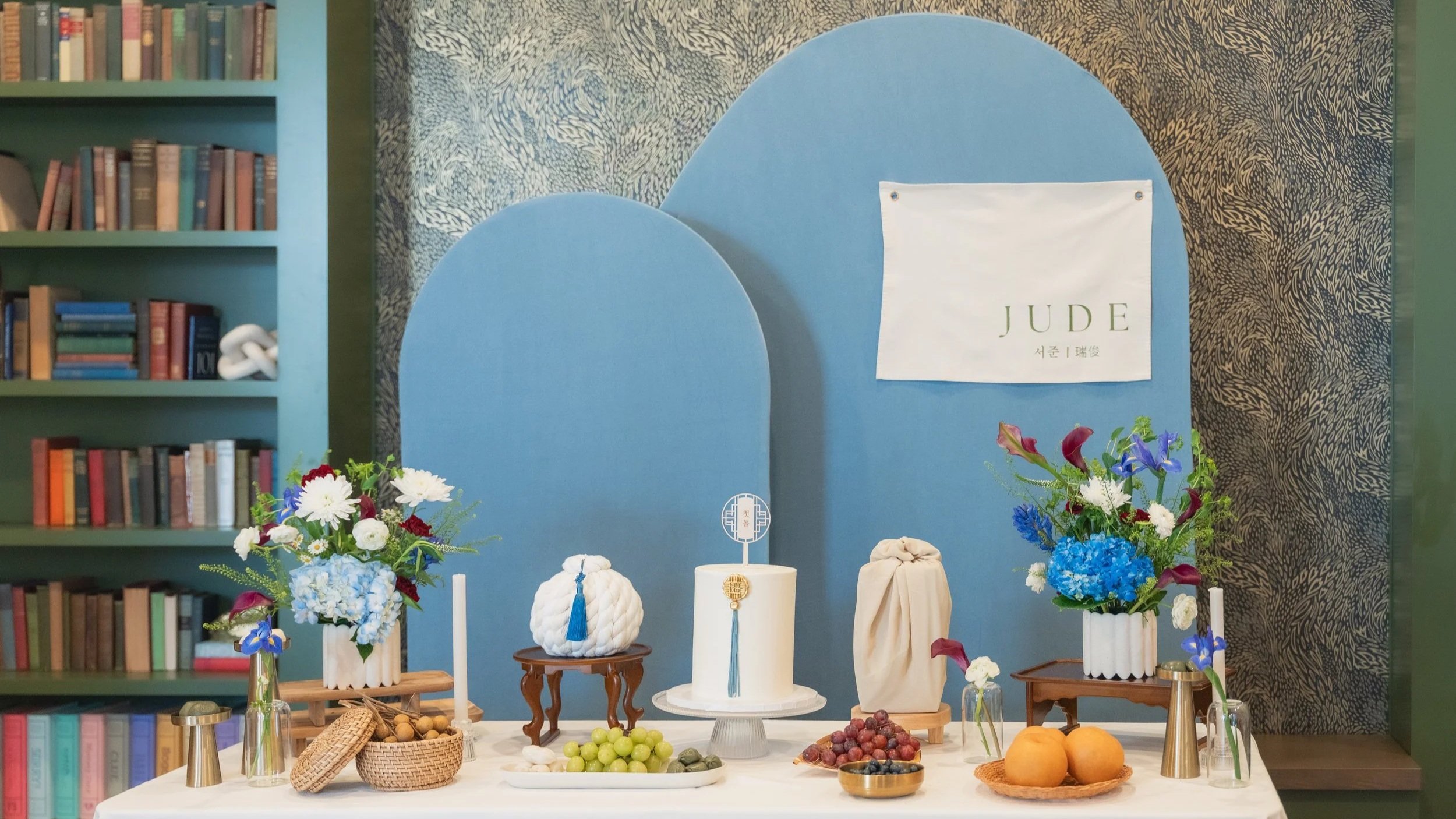 A decorated table with a cake, various fruits, and flowers, set against a backdrop with blue arches and bookshelves on either side.