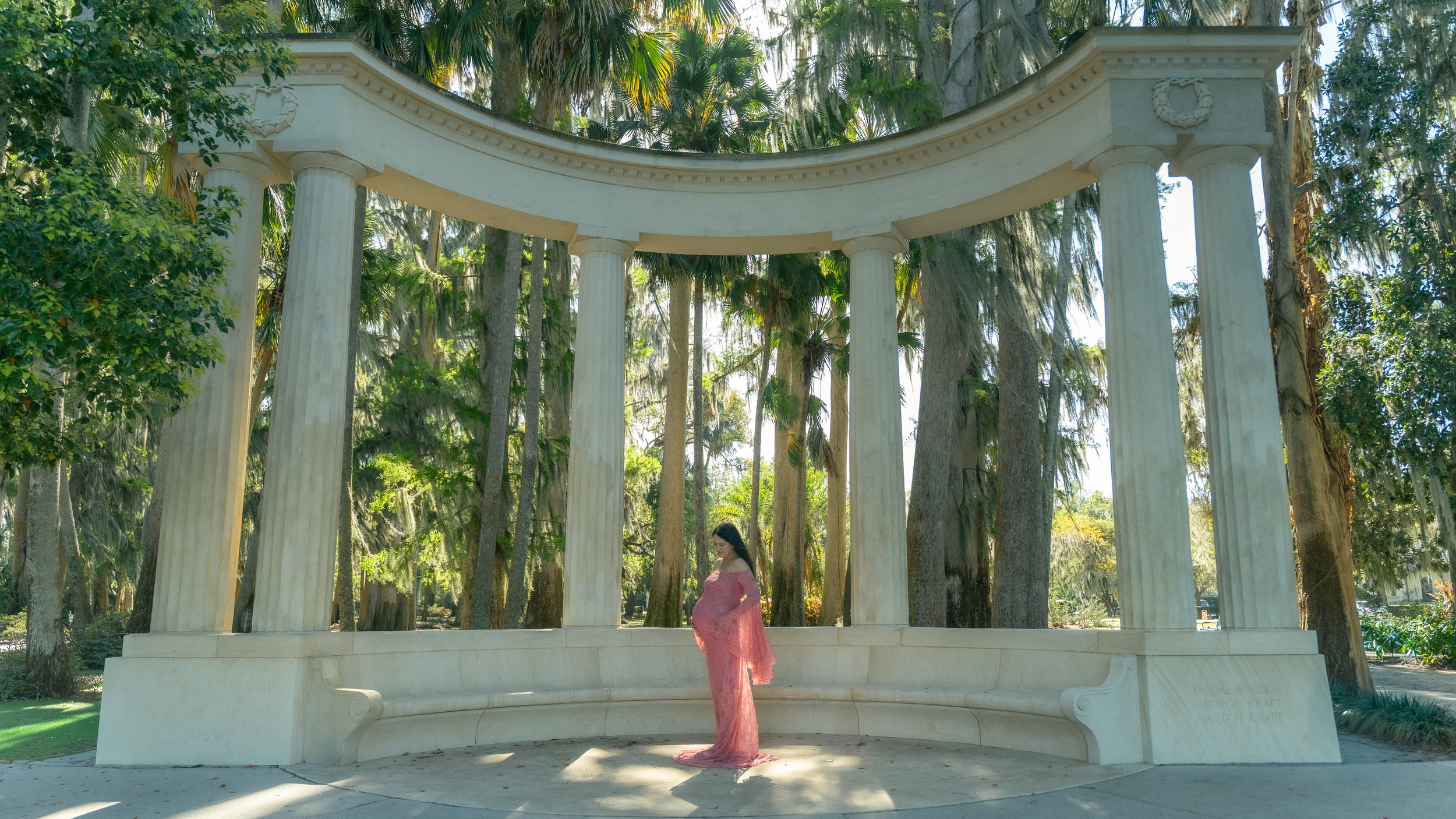 A pregnant woman in a pink dress standing under a white classical bandstand with columns in a park surrounded by tall trees for a maternity photography session.