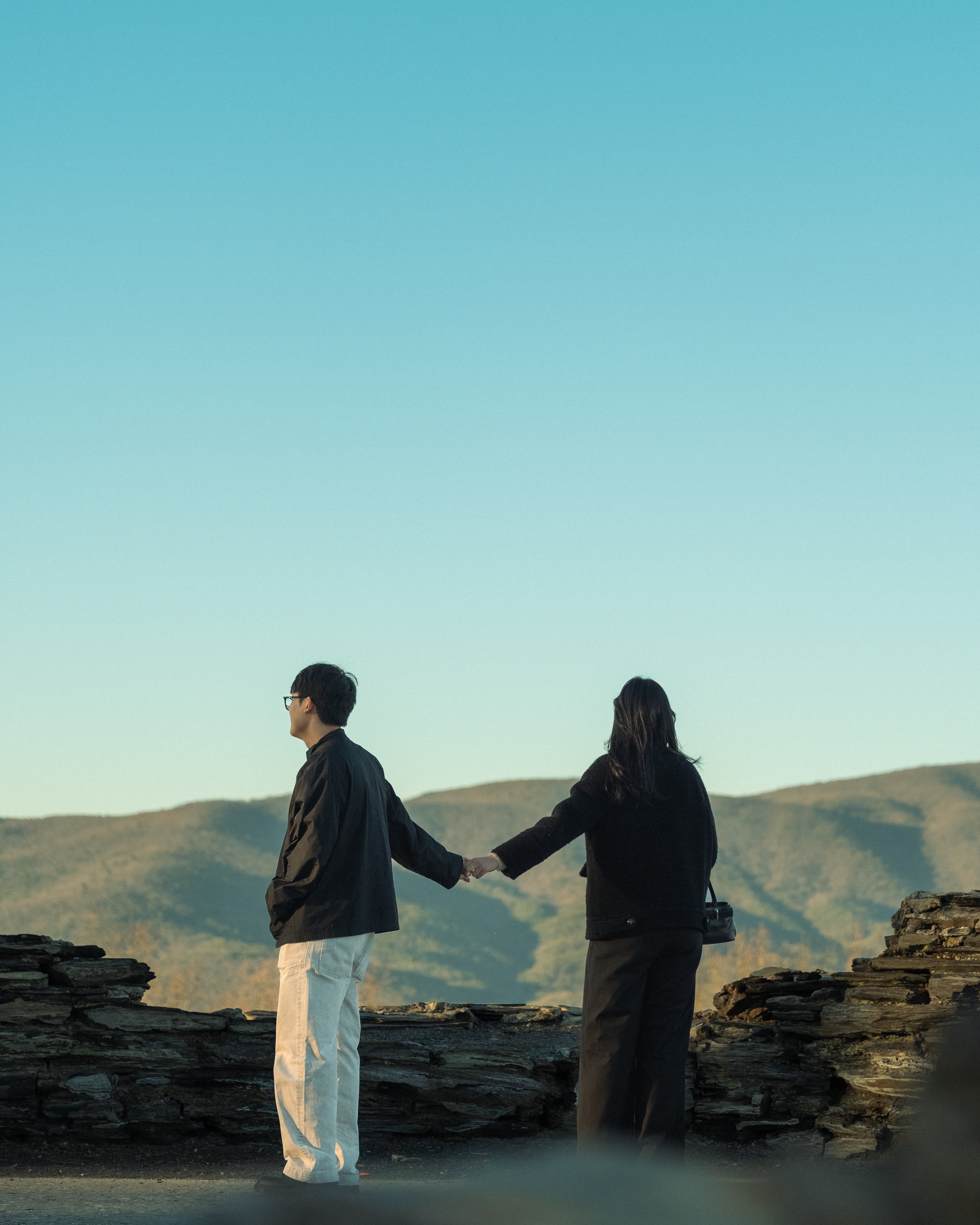 Two people holding hands outdoors, standing on rocky terrain, with a mountain range in the background during sunset or sunrise.