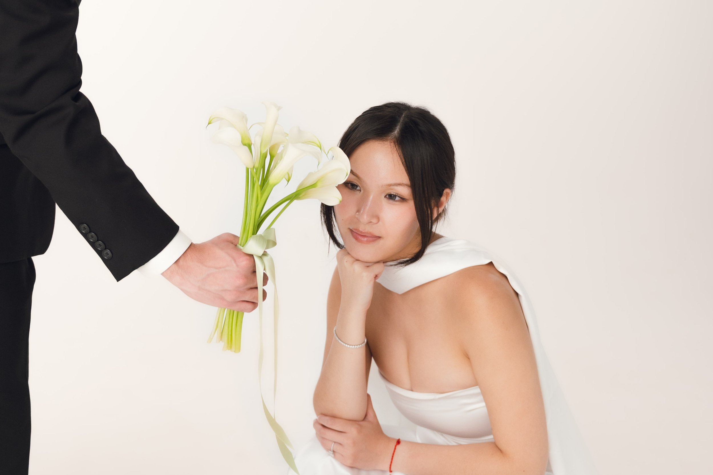 A woman with dark hair gazes thoughtfully at white calla lilies being handed to her by someone wearing a black suit, against a plain white background.