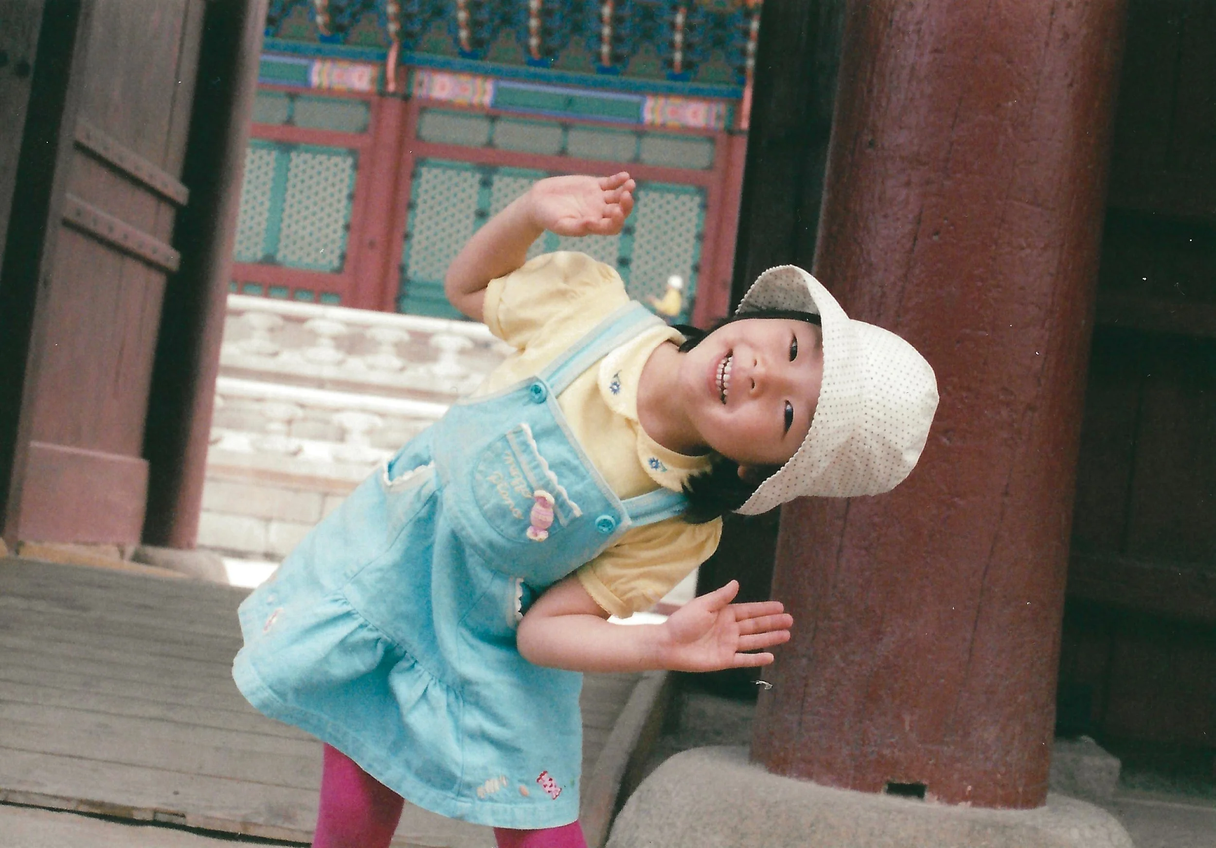 A young girl smiling and clapping with her hands in a praying position, wearing a white hat, yellow shirt, and blue overalls, standing under a wooden structure with colorful background details.