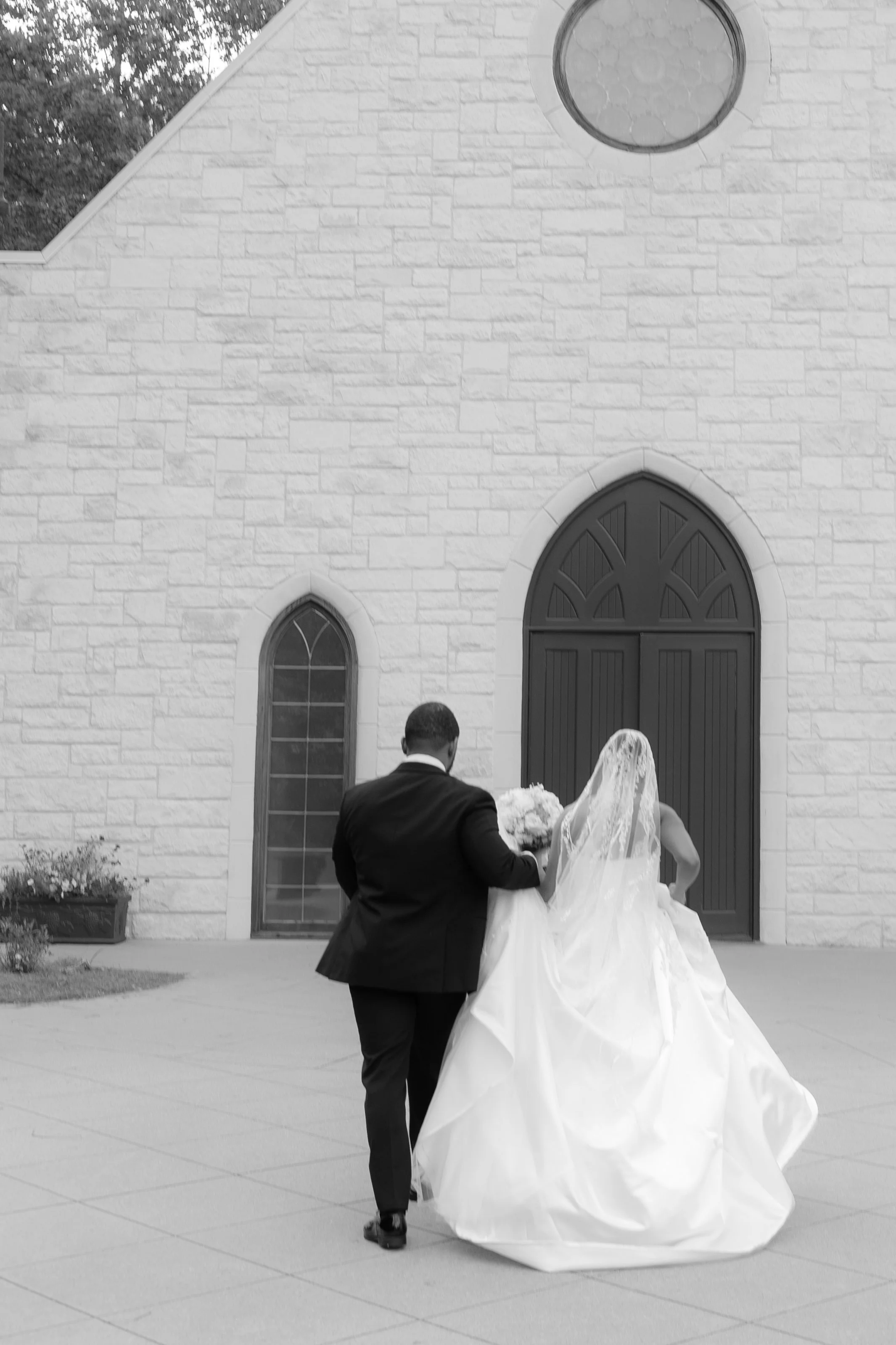 A bride and groom walking out of a church, with the bride wearing a wedding gown with a veil and holding a bouquet, and the groom wearing a suit, as they leave a building with a brick facade and arched doors.