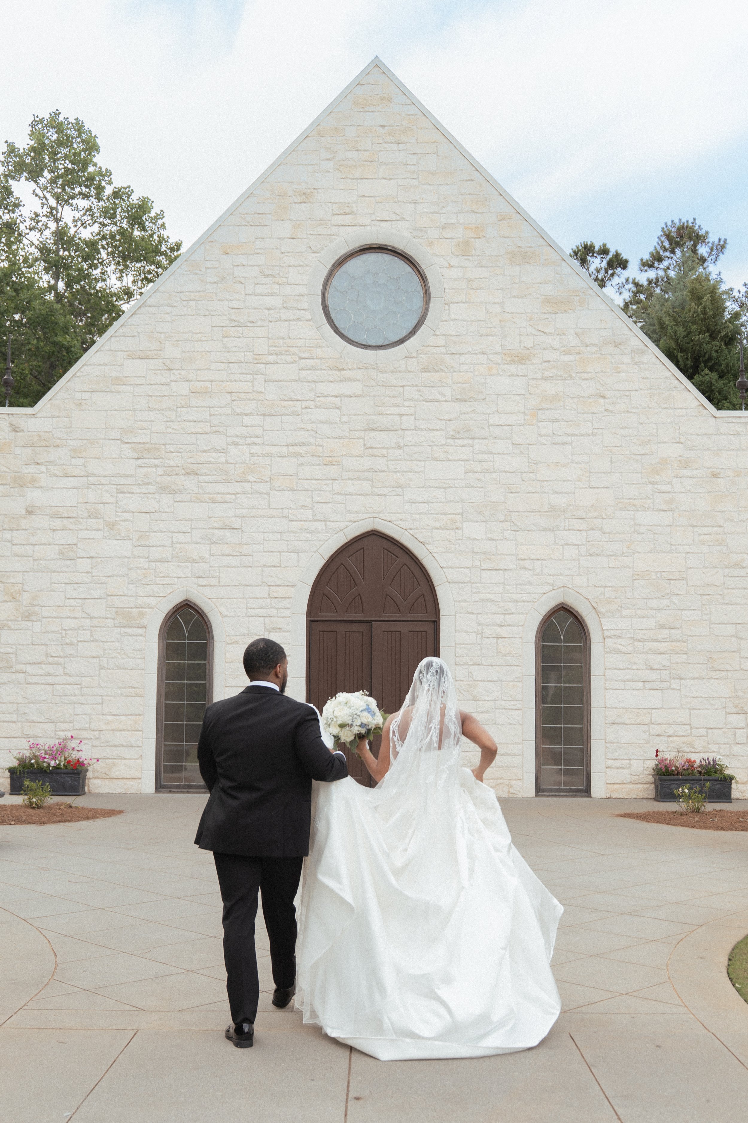 A bride and groom walking away from the camera outside a church, holding hands. The groom is in a black suit and the bride is in a white wedding dress with a veil. The church has a beige stone facade with arched windows and a circular window above the entrance.