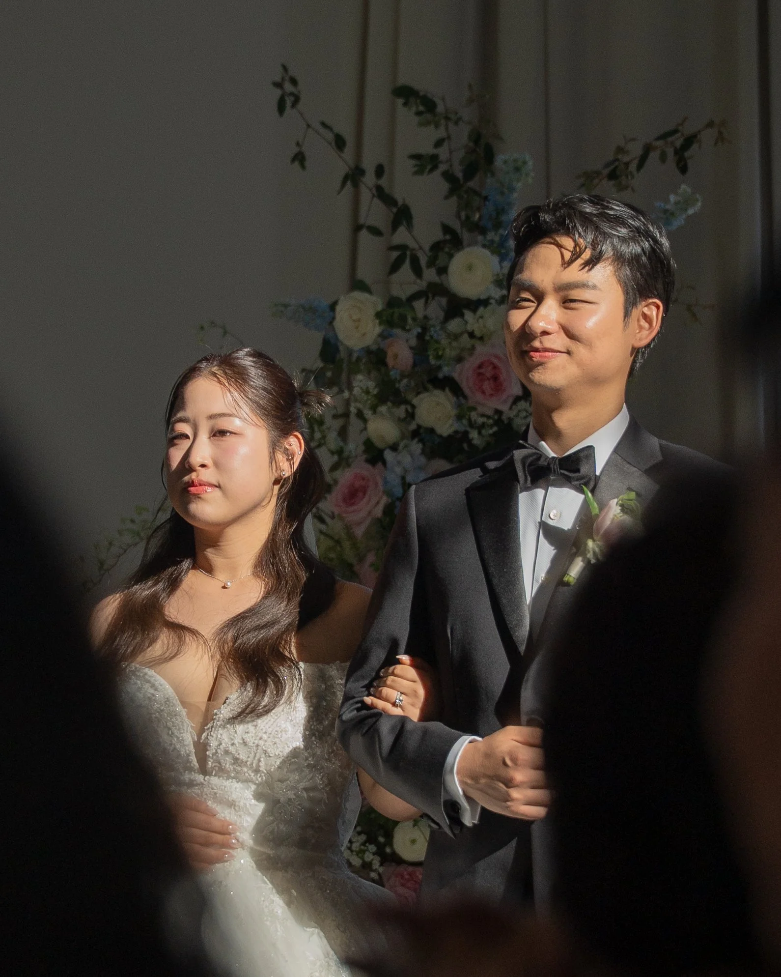 A bride and groom standing together during their wedding ceremony, with a floral backdrop behind them. The bride is wearing a lace wedding dress and a necklace, with her hair styled in waves. The groom is dressed in a tuxedo with a bow tie and boutonniere, holding the bride's hand. Both are smiling softly.
