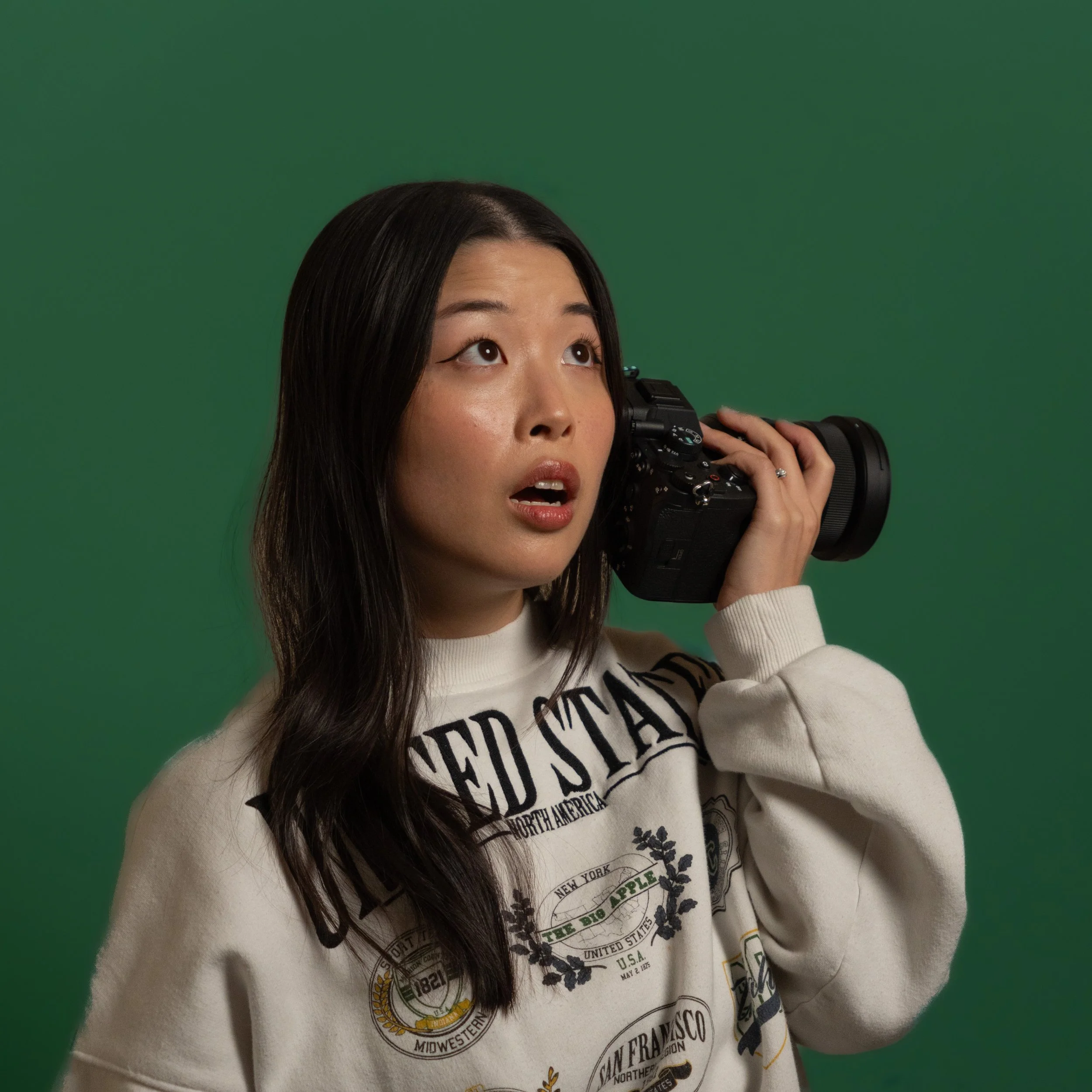 A woman with long dark hair looks upward while holding a camera to her face, set against a plain green background.