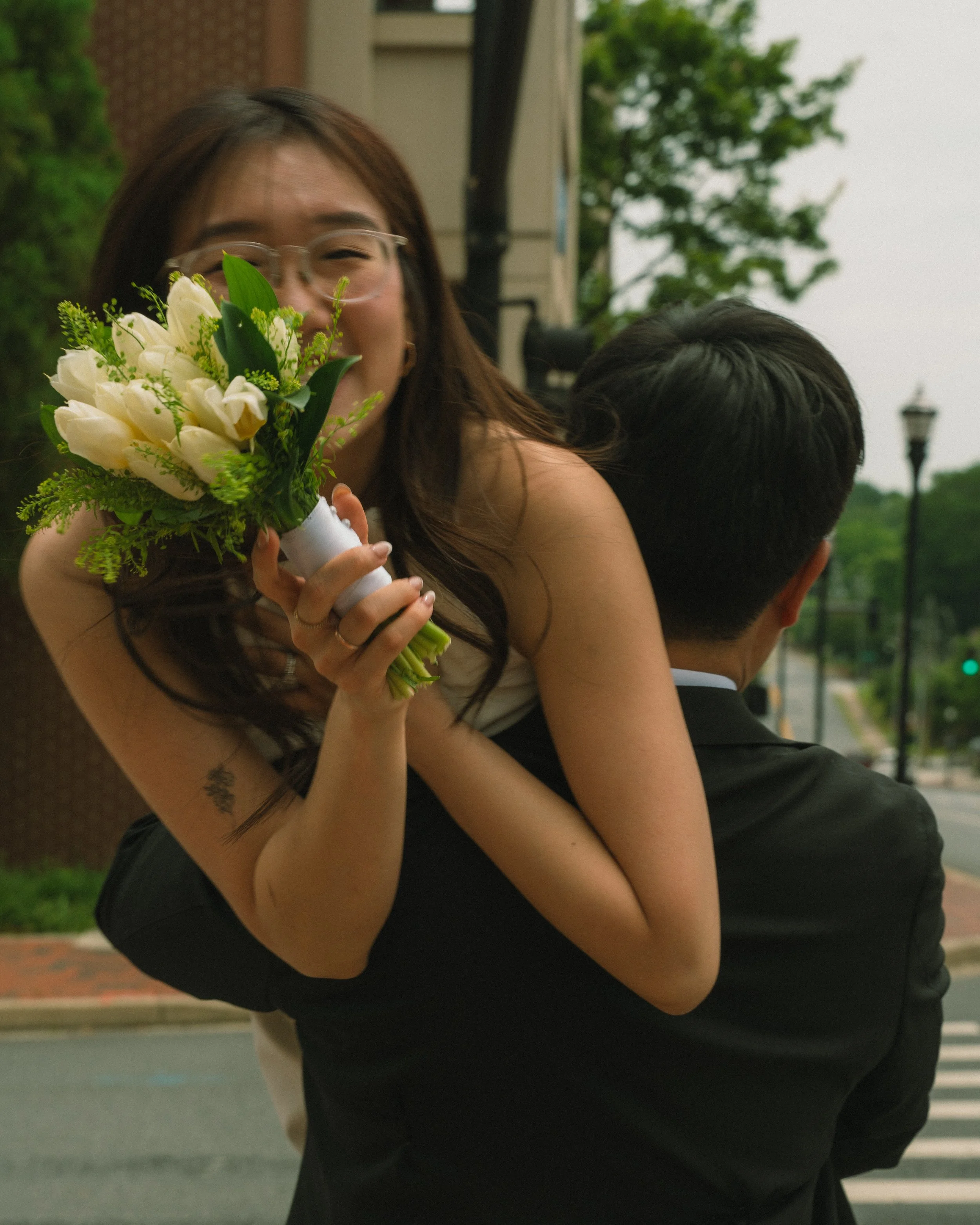 A young woman with glasses, holding a bouquet of white lilies and greenery, smiling and embracing someone outdoors on a cloudy day.