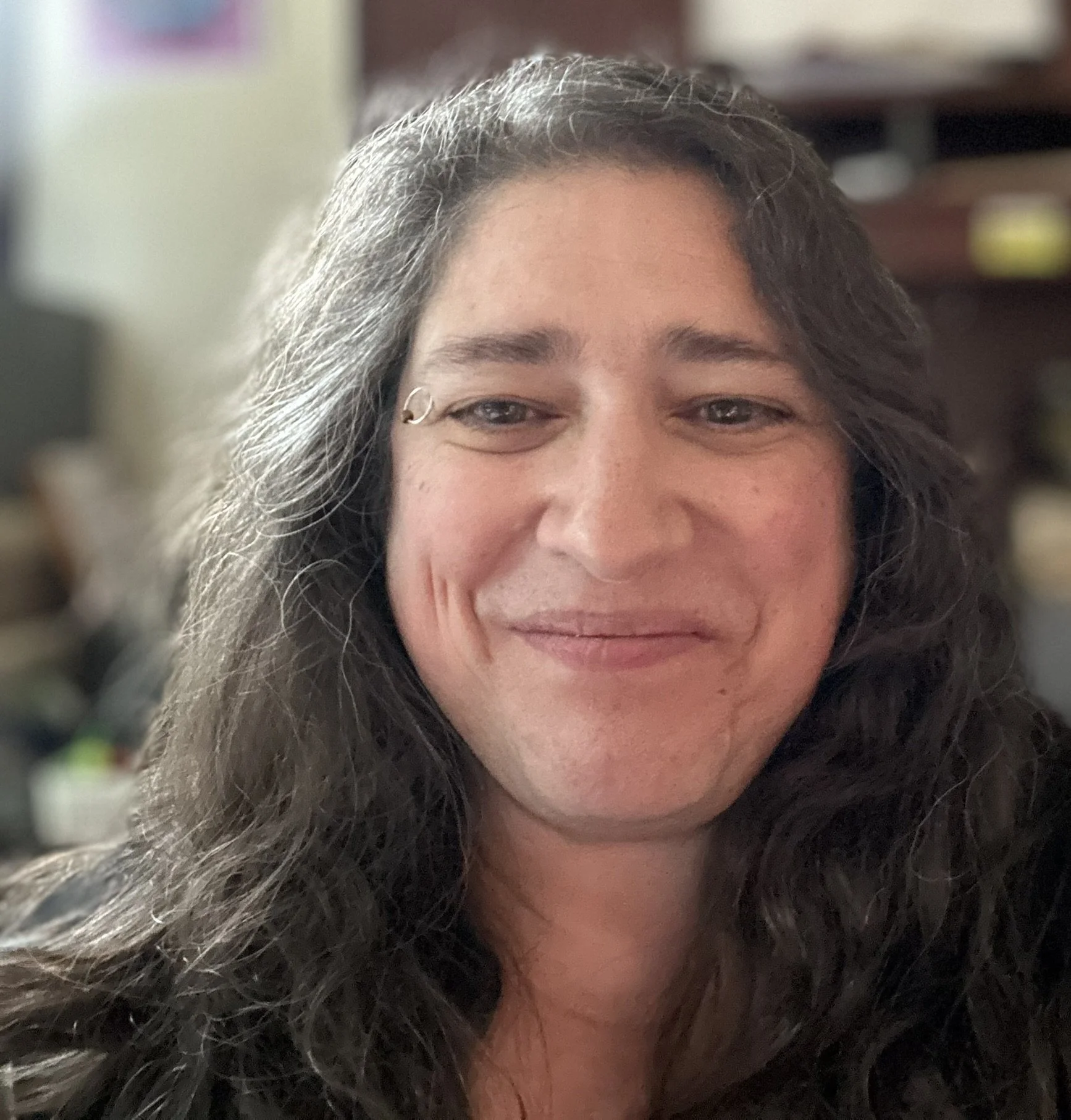 Close-up of a smiling woman's face with long gray hair, indoors with shelves in the background.