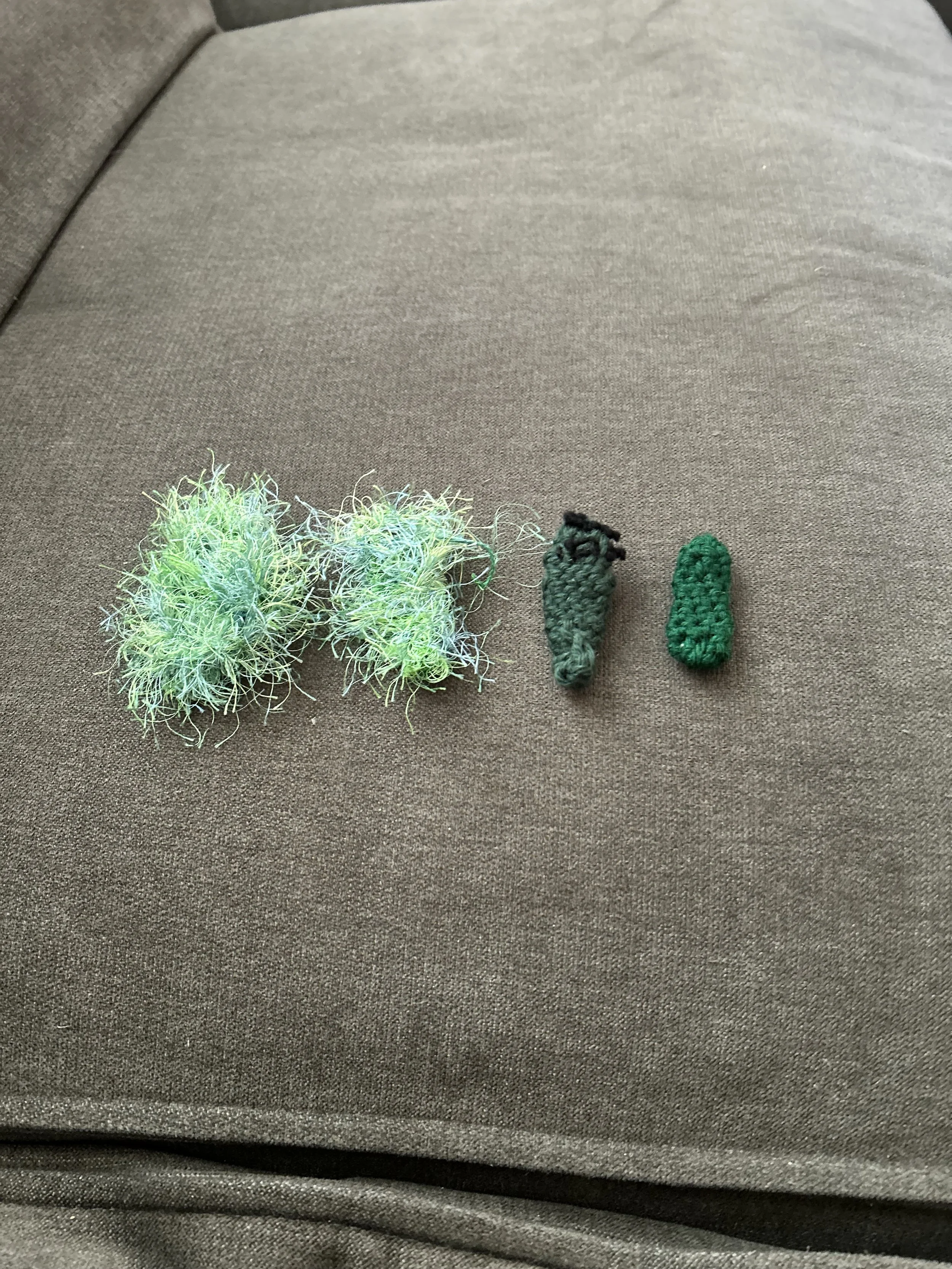 Close-up of a gray fabric surface with three small crocheted objects, two green and one gray, resembling yarn pieces or small toys.
