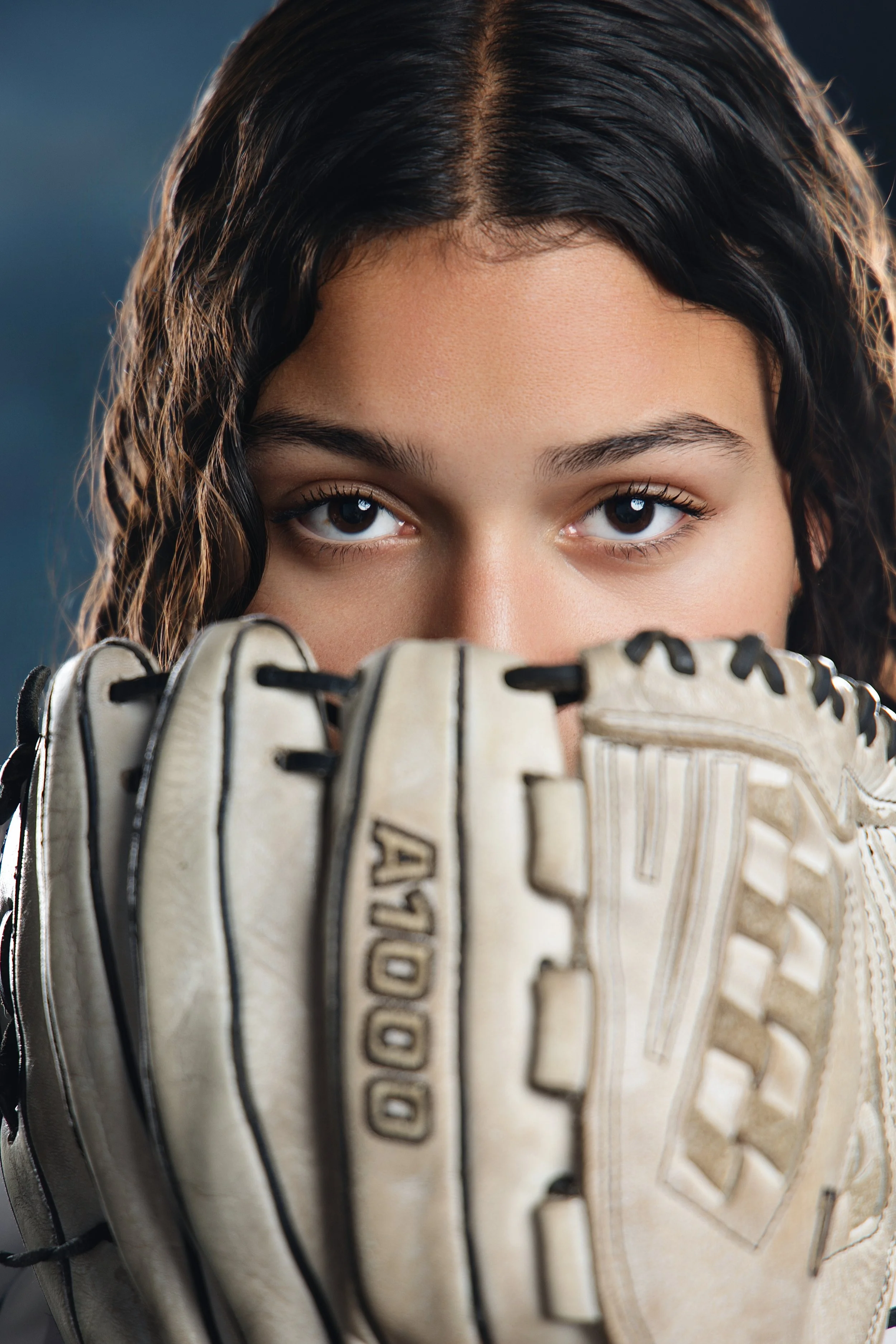 Close-up of a young woman's face, partially obscured by a baseball glove. She has dark wavy hair, and her brown eyes are visible as she looks directly at the camera.