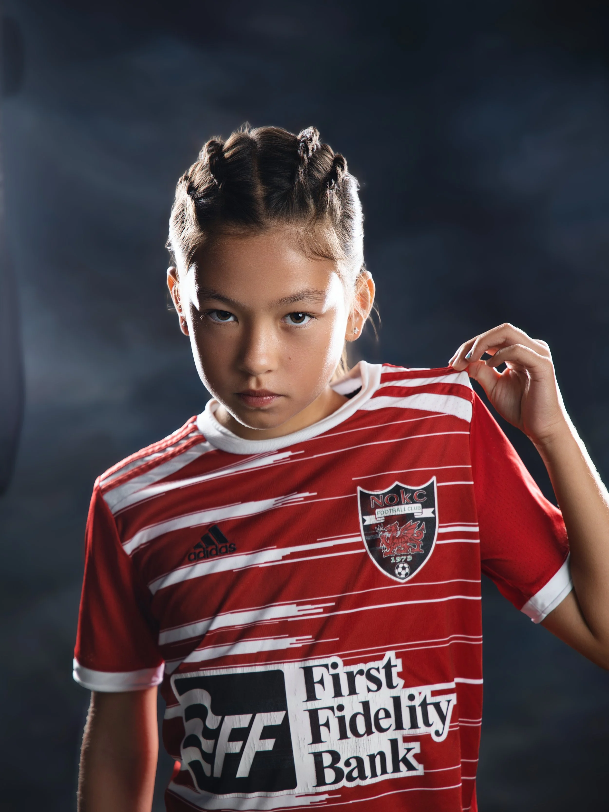 A young girl with braided hair wearing a red and white striped sports jersey, holding her collar with her left hand, in front of a dark background.