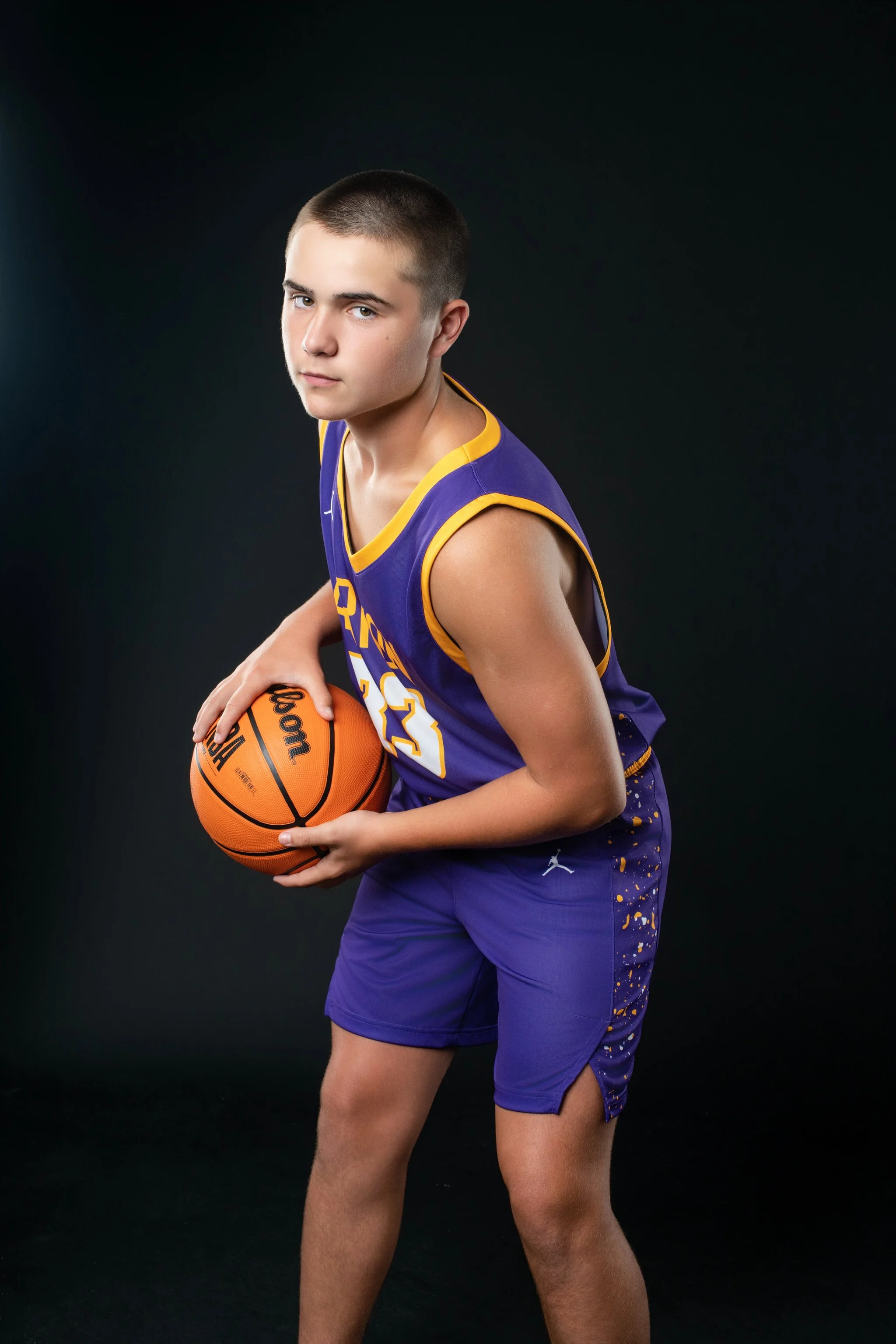 Young male basketball player in purple uniform holding a basketball against a dark background.