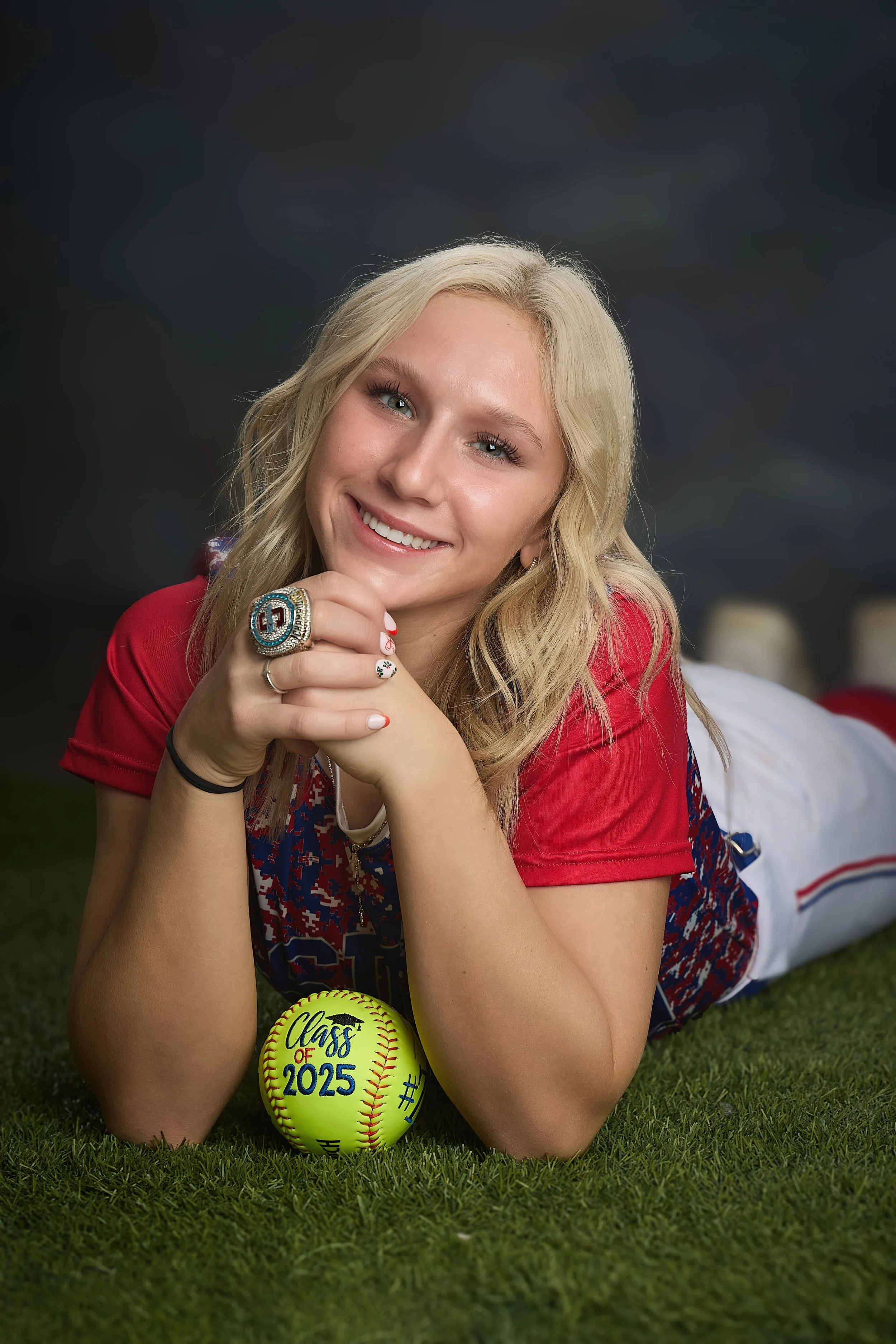 A young woman lying on grass, smiling, with blonde hair, wearing a red and blue patterned shirt, a large ring, and sports-themed nail polish. She is resting her chin on her hands next to a yellow softball that has 