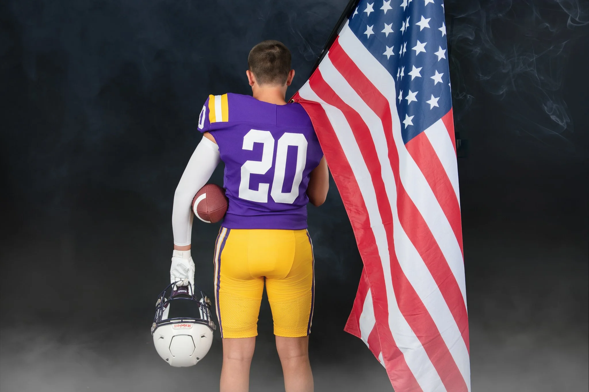 A person wearing purple and yellow sportswear holding a football and a helmet, standing with their back to the camera, holding an American flag, against a dark background with smoke.