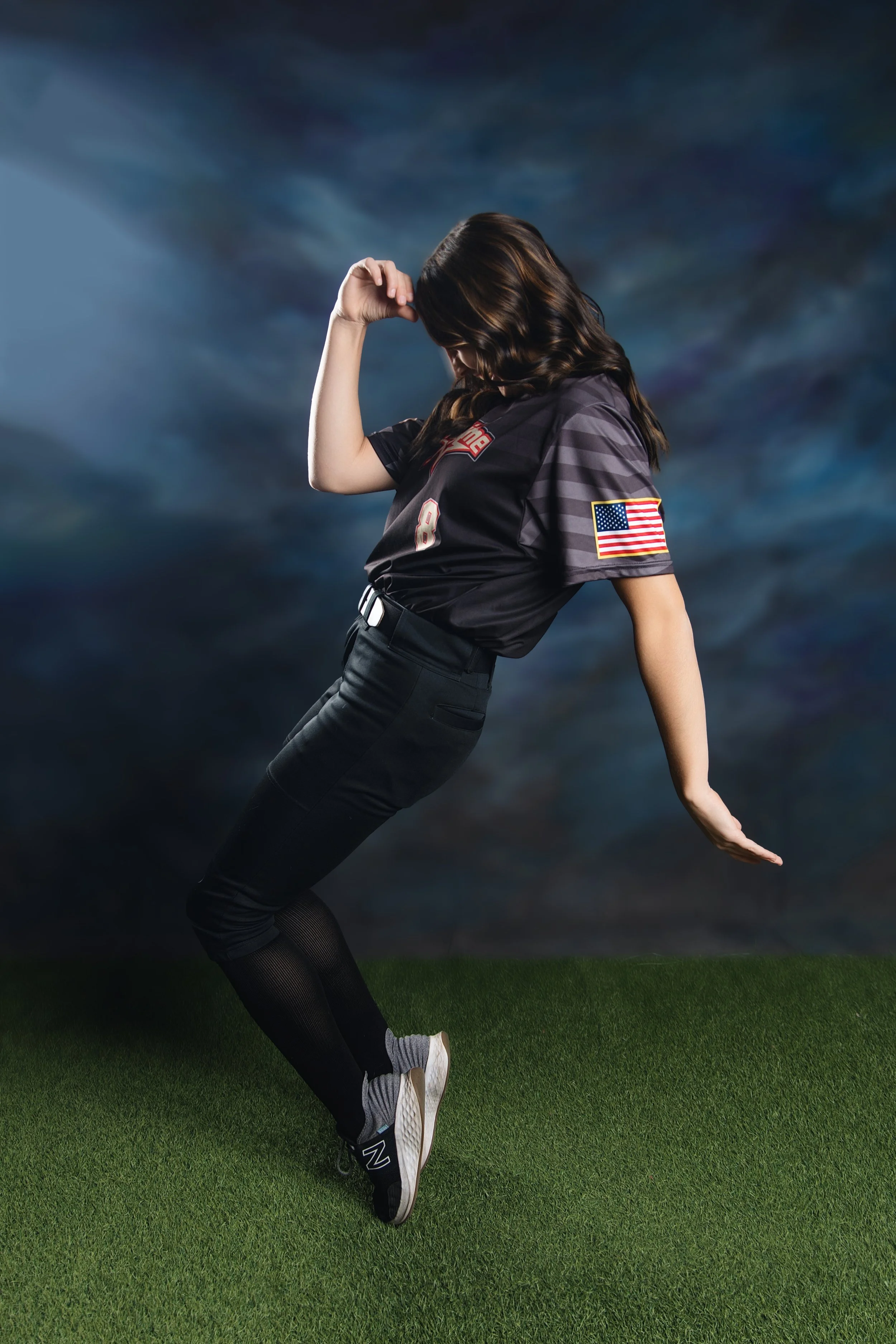 A woman wearing a black sports jersey with an American flag patch on the sleeve, black pants, and sneakers, is posing on a grassy field against a dark, cloudy sky background. She appears to be balancing on her left leg with her right leg bent and her