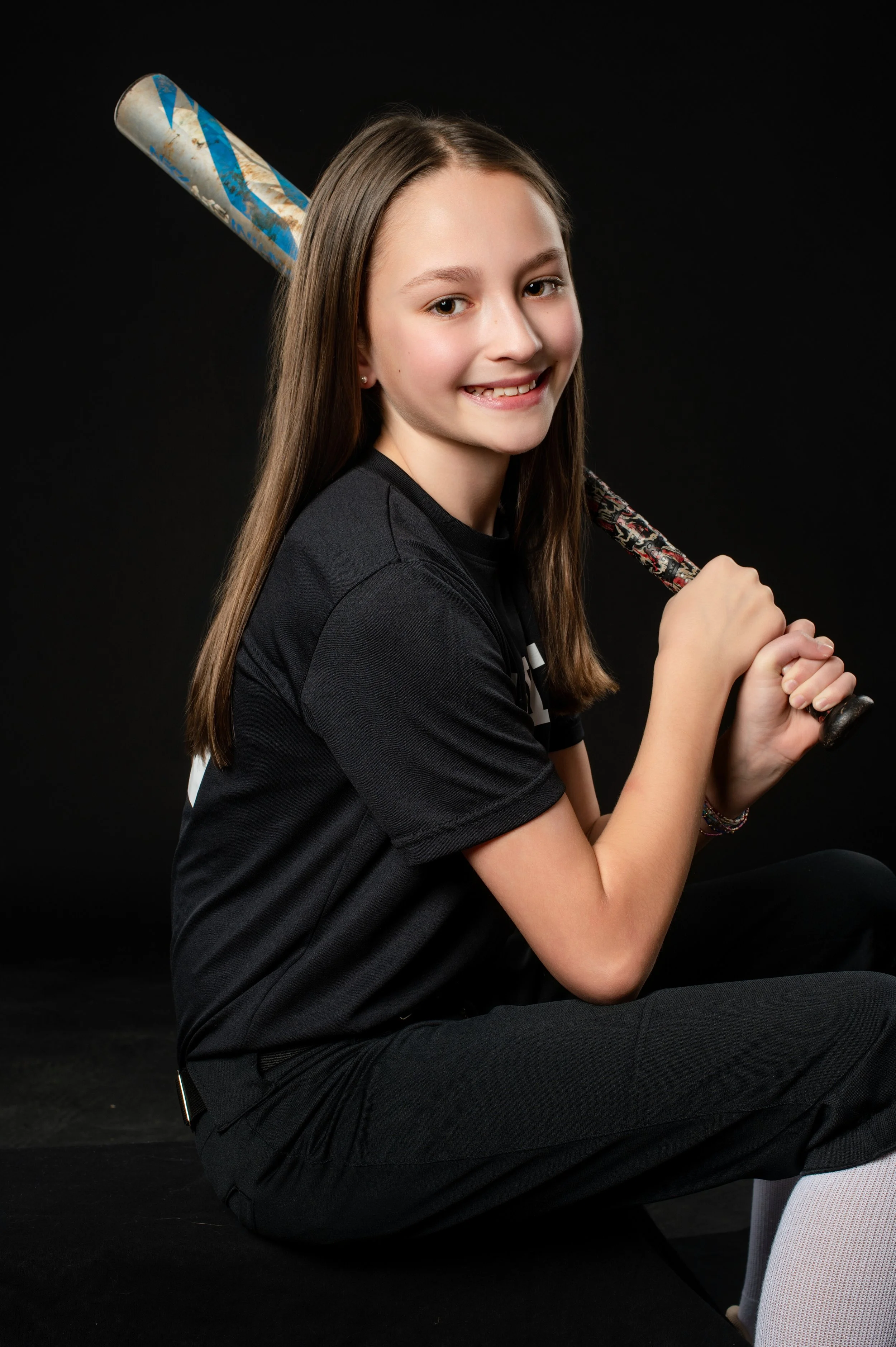 Girl sitting on the floor with a baseball bat over her shoulder, smiling, wearing a black shirt and black pants, against a black background.