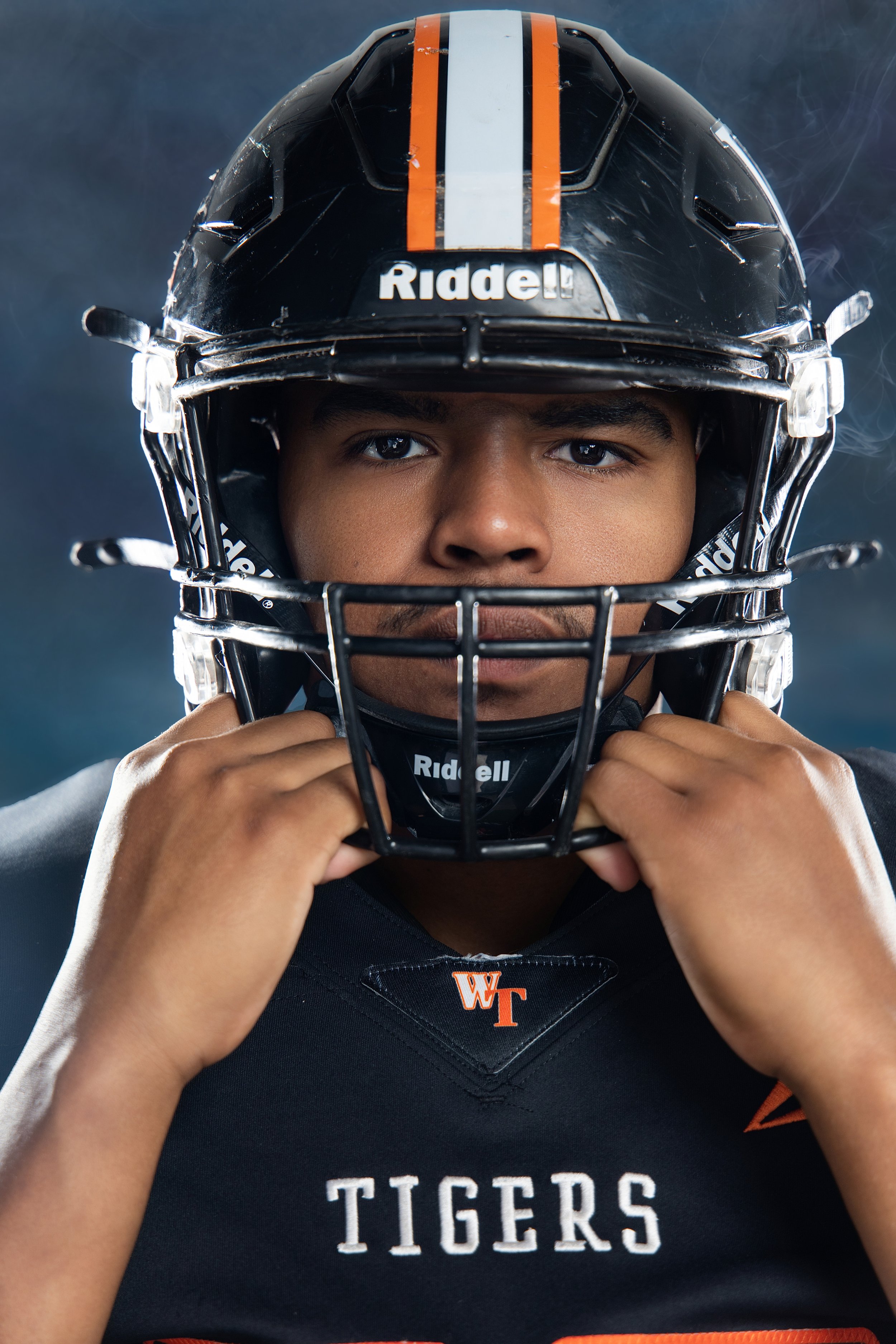 A young person wearing a black football helmet with orange and white stripes, holding the chin strap with both hands. Their face is partially visible, with dark eyes and skin. They are also wearing a black football jersey with the words "Tigers" visi