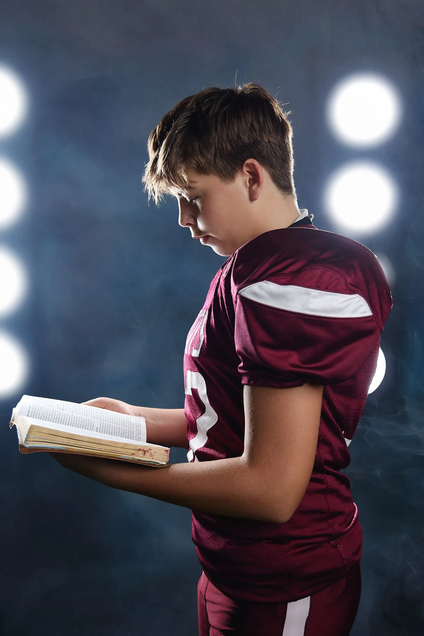 Young male football player in a maroon jersey with white details, holding and reading a book, with bright stadium lights in the background.