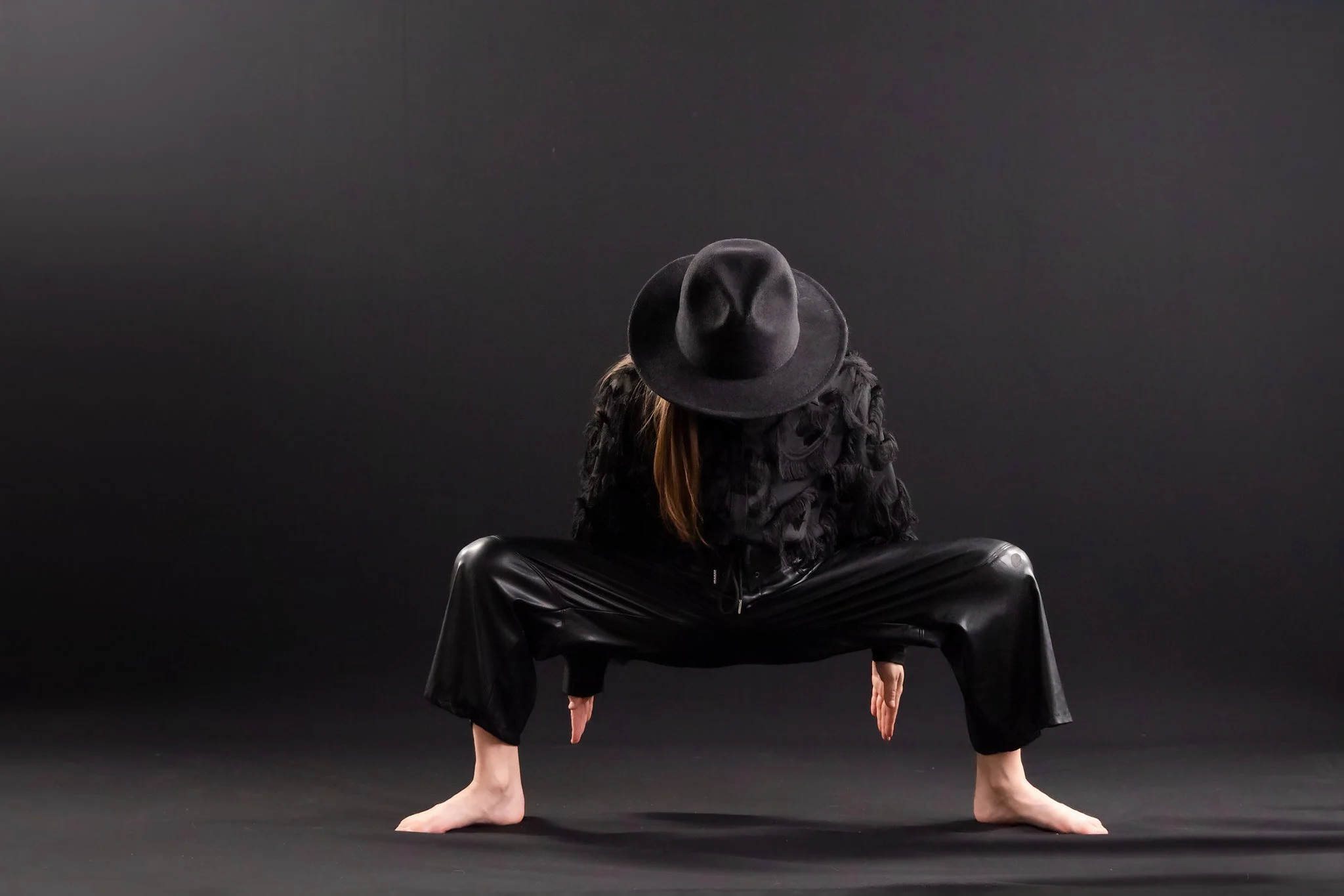 A person with long hair, wearing a large black hat, black fur scarf, black jacket, and black pants, is crouching with hands and feet on the ground against a dark background.