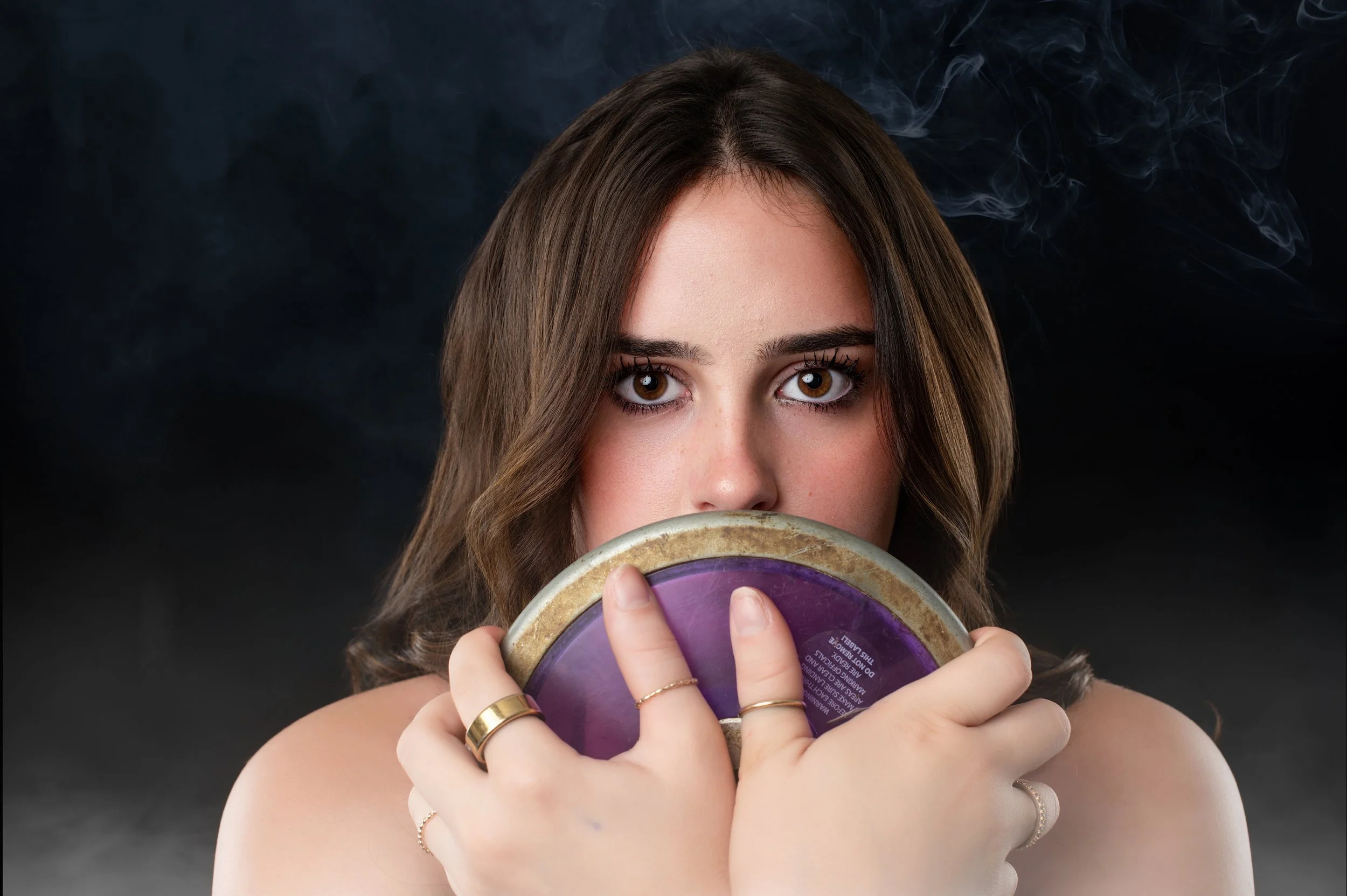 Young woman with brown hair and brown eyes holding a purple and beige painted ceramic bowl in front of her face, with her lips visible behind the bowl, against a dark background with smoke or mist.