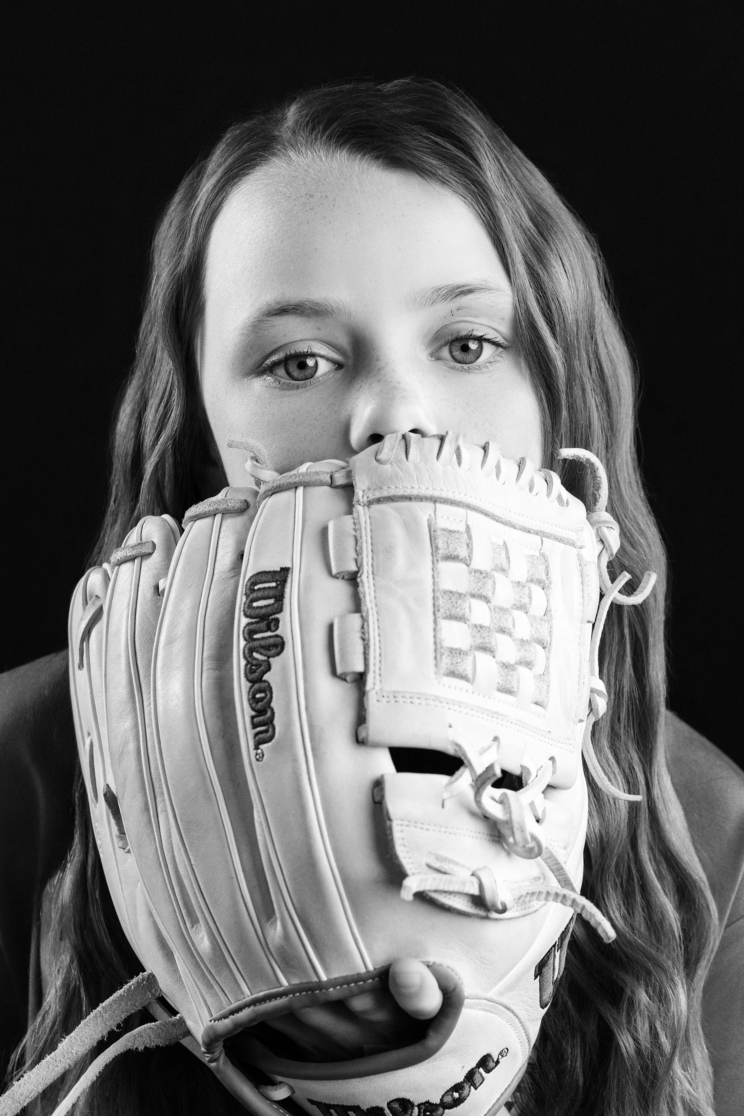 A young girl with long hair holds a baseball glove up to her face, partially covering her nose and mouth, with only her eyes and forehead visible, against a black background.