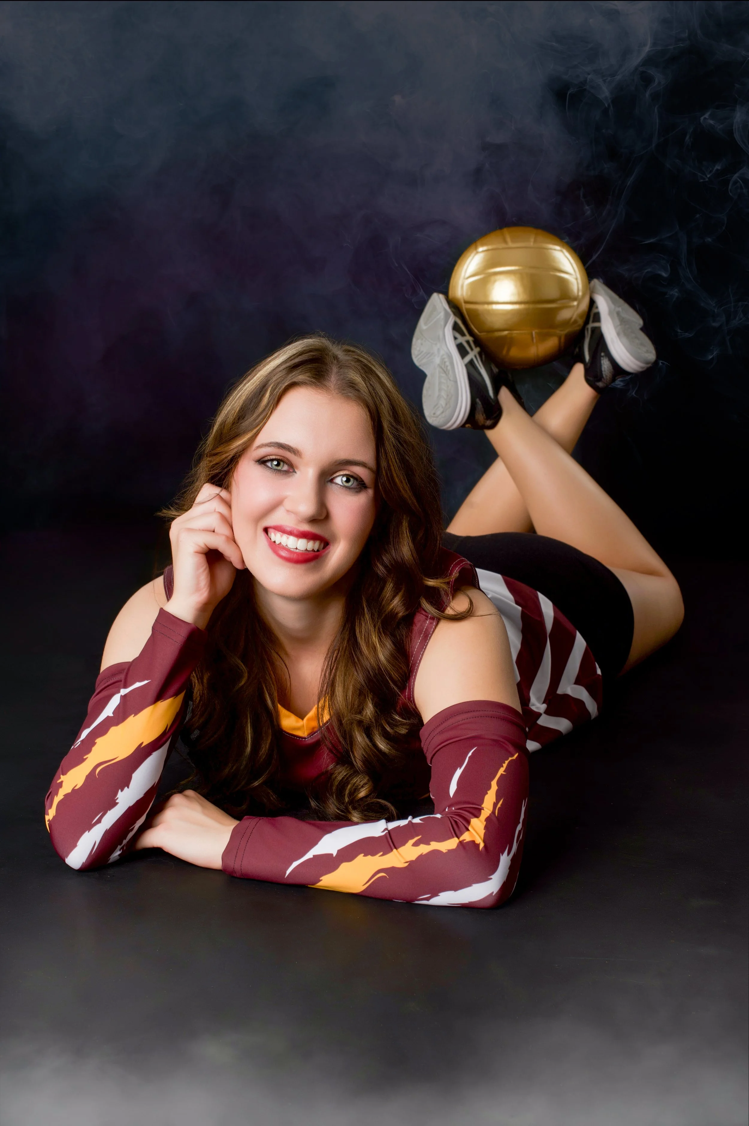 A young woman with long brown hair and makeup, smiling, laying on her stomach with her head resting on her hand, wearing a long-sleeved sports jersey with maroon, yellow, and white colors, and football shoes, with a gold football ball elevated on her