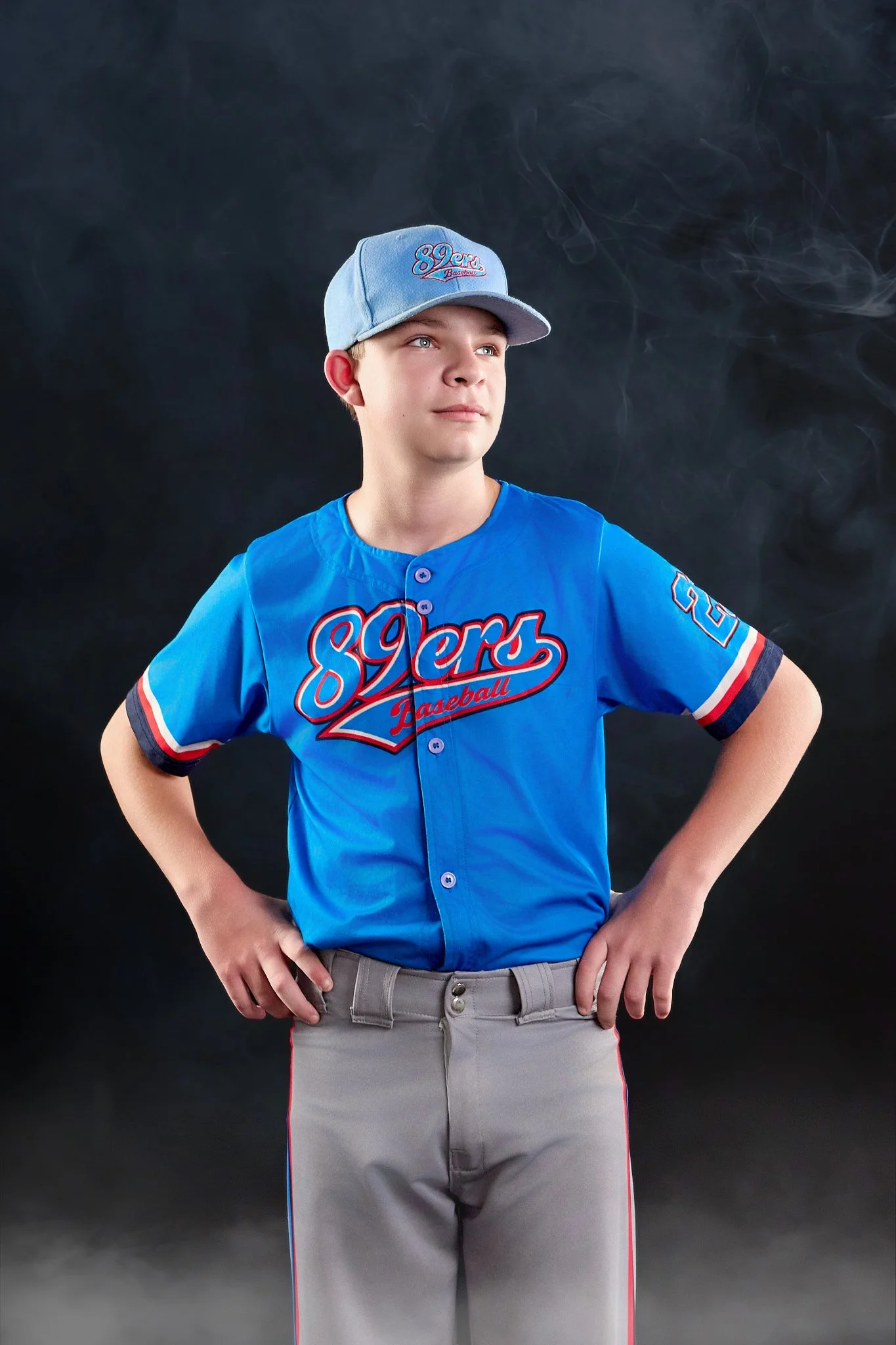 A young boy wearing a blue baseball uniform with red and white accents, a gray and blue cap with '89ers Baseball' embroidered on it, standing with hands on hips against a dark smoky background.