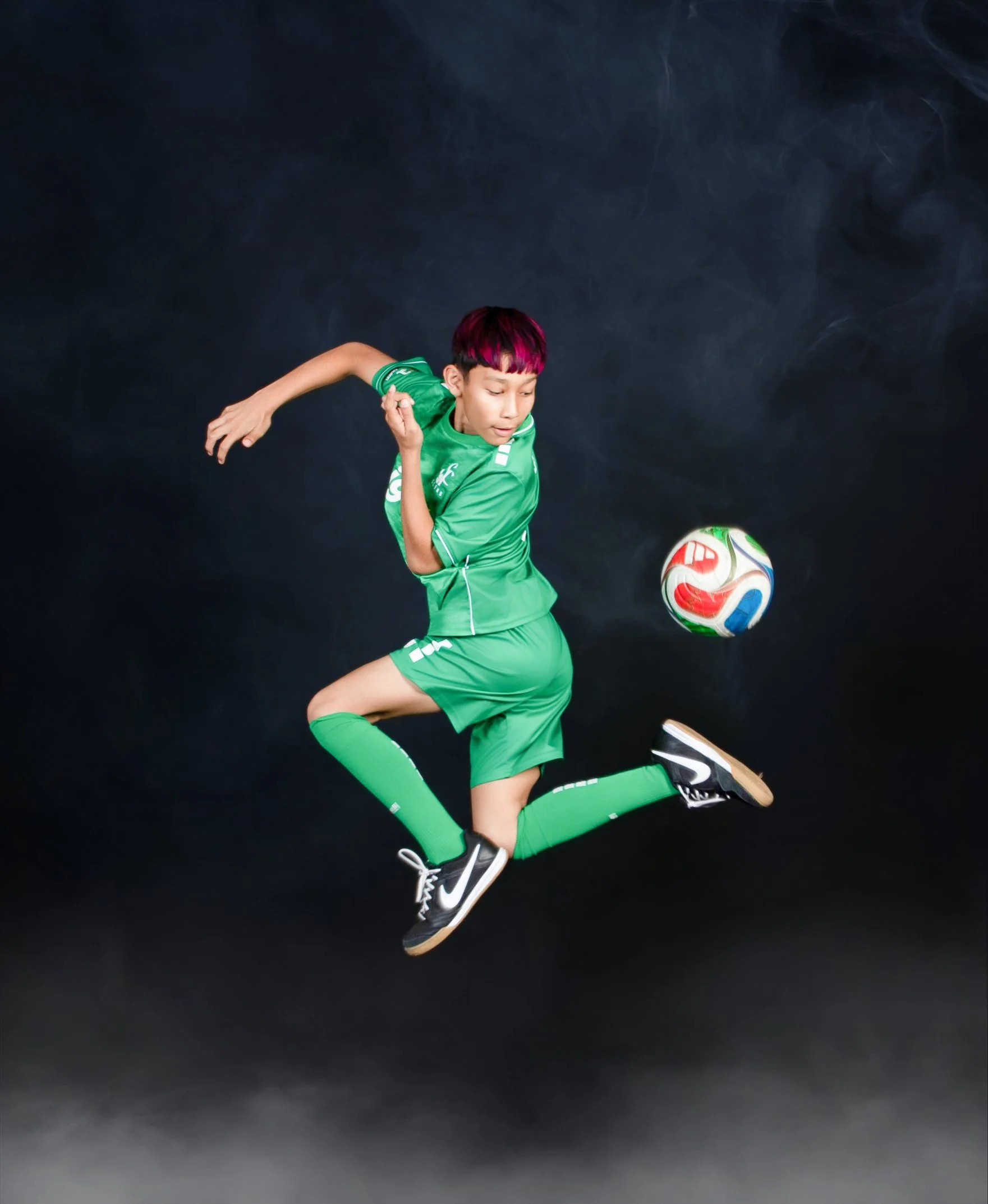 A young boy wearing a green soccer uniform and black sneakers is captured mid-air as he kicks a colorful soccer ball against a dark background.